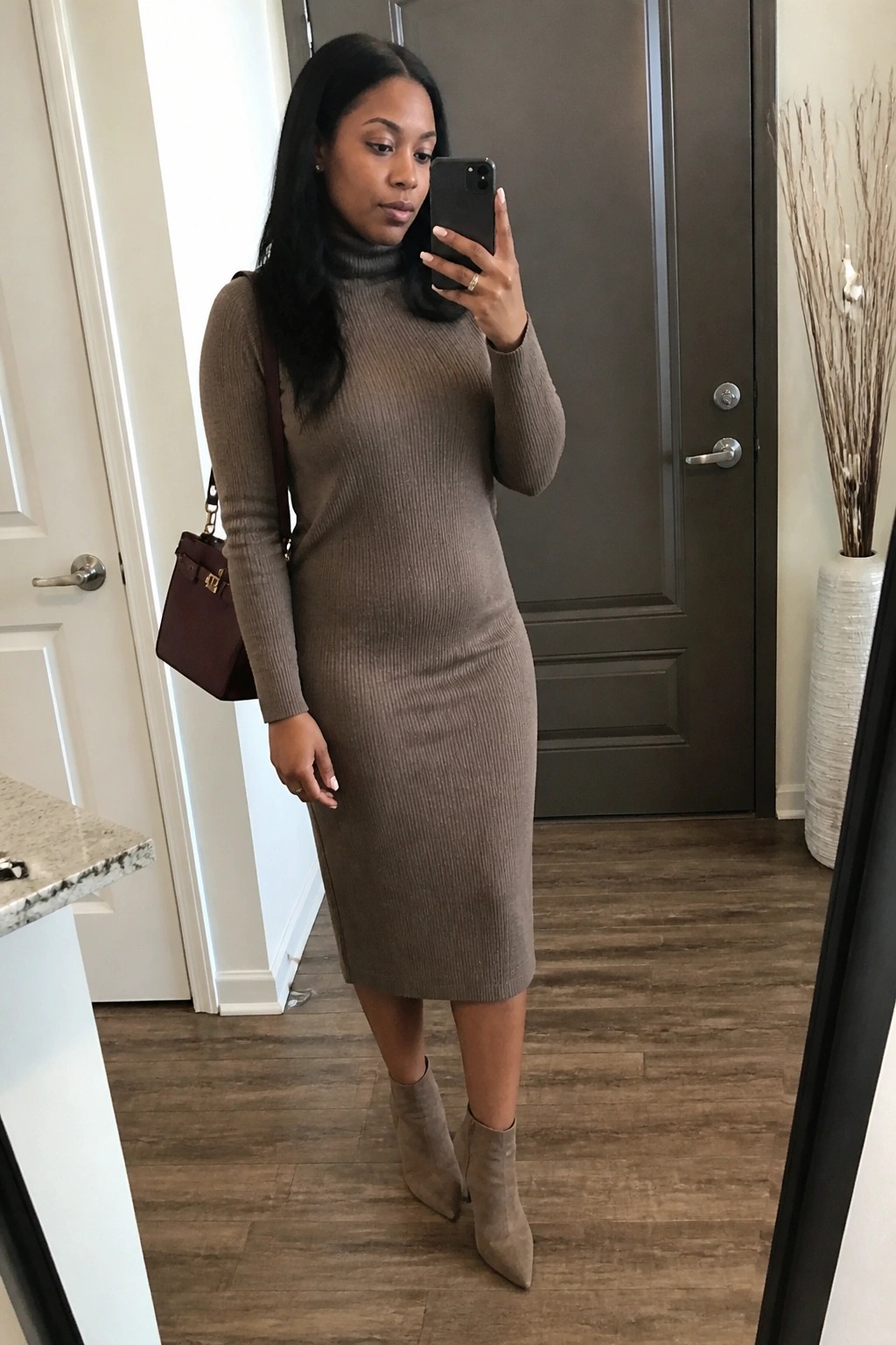 Woman in taupe turtleneck sweater dress and beige boots taking mirror selfie with maroon bag