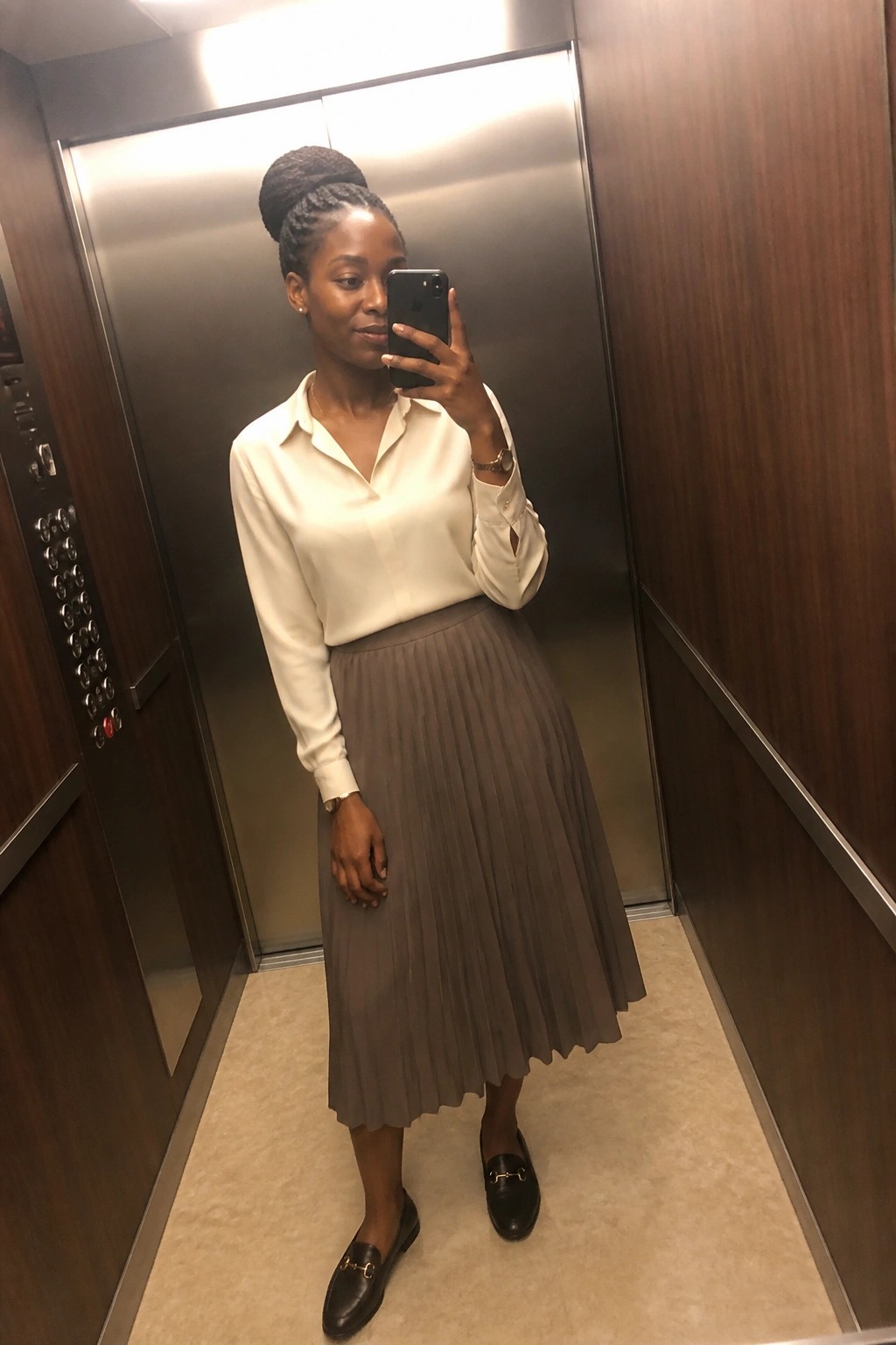 Woman in white blouse and taupe pleated skirt taking selfie in elevator