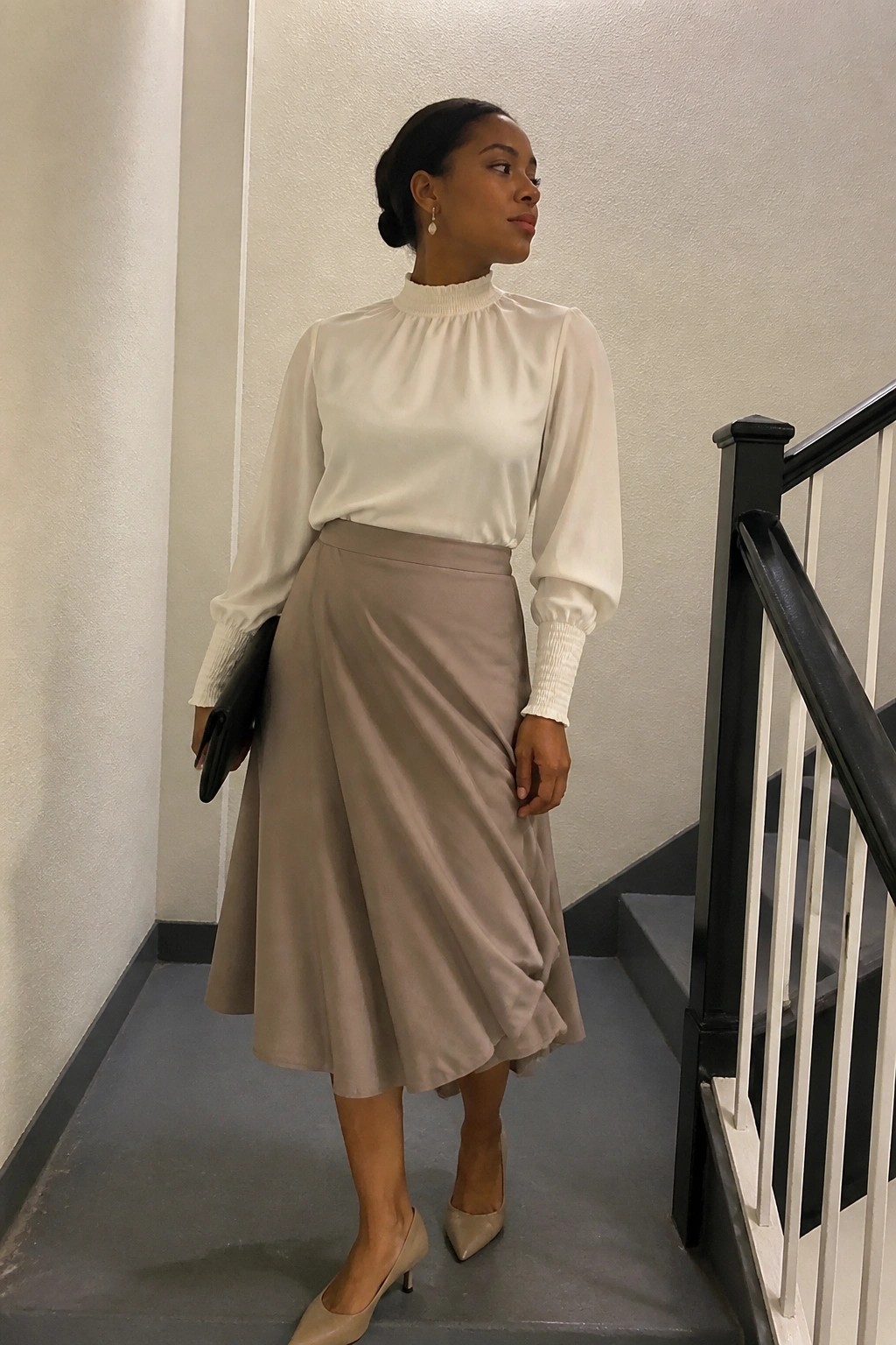 Woman in white blouse and beige skirt holding black clutch on stairs