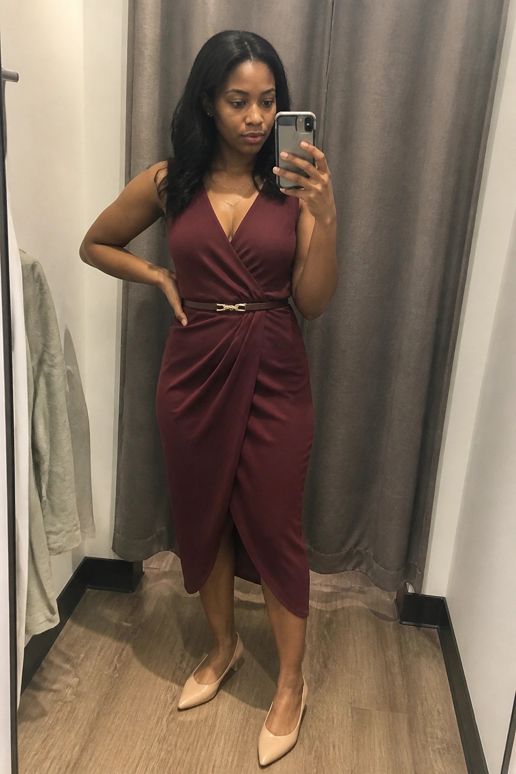 Woman in maroon wrap dress taking mirror selfie in fitting room with beige heels.