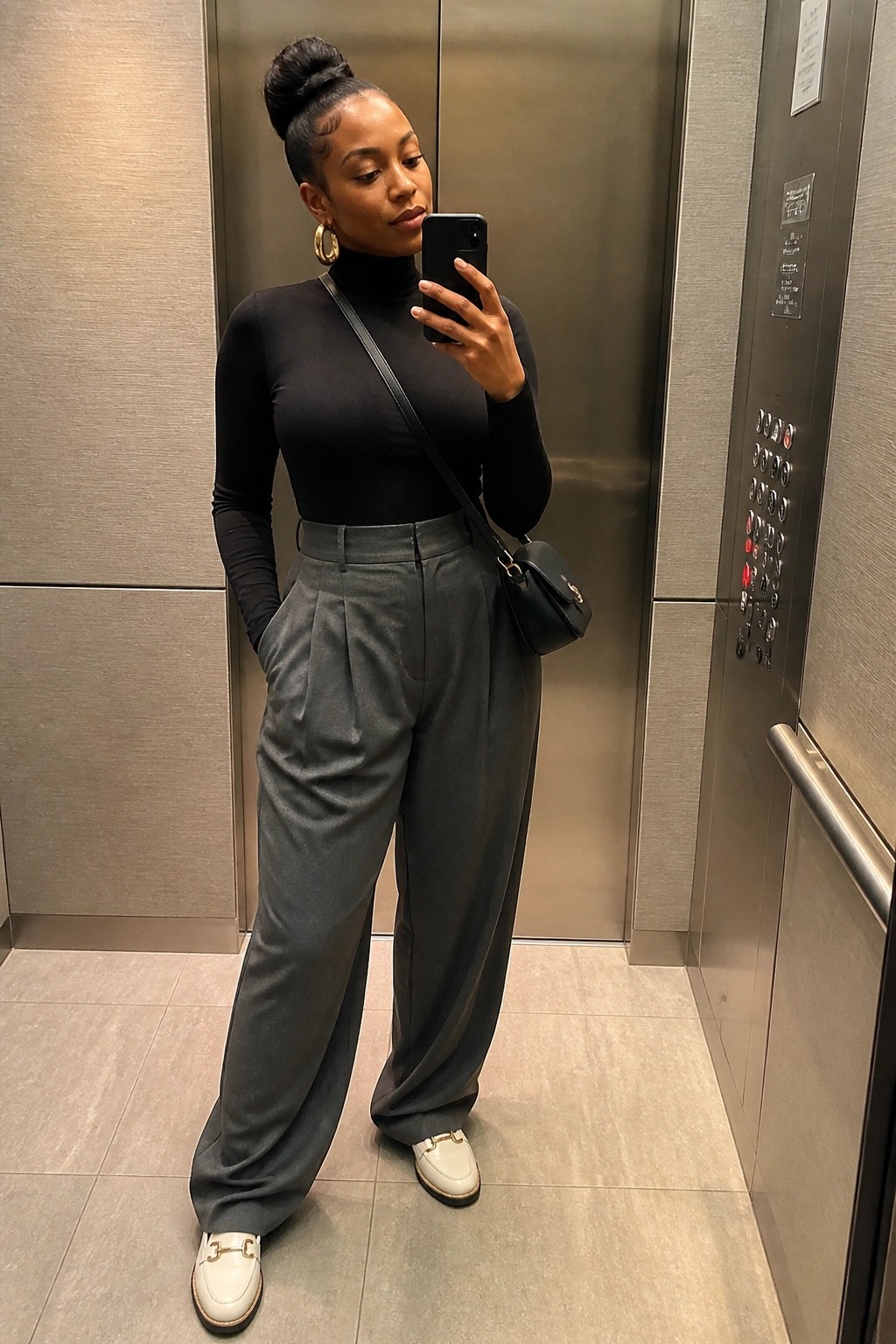 Woman in black turtleneck and gray trousers taking selfie in elevator