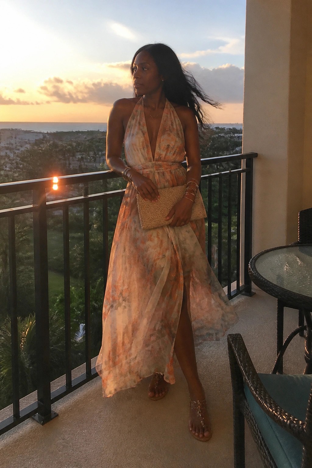 Woman wearing a flowing halter maxi dress with watercolor orange and beige print, deep V neckline, side slit, paired with strappy sandals, woven clutch, layered bracelets, and delicate necklace.