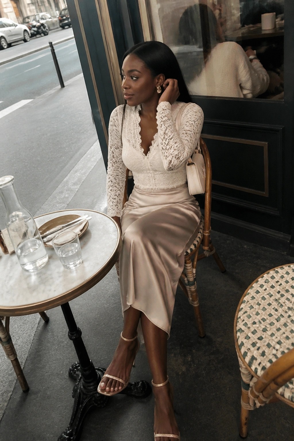 Stylish woman in white lace top and beige satin skirt at sidewalk cafe.