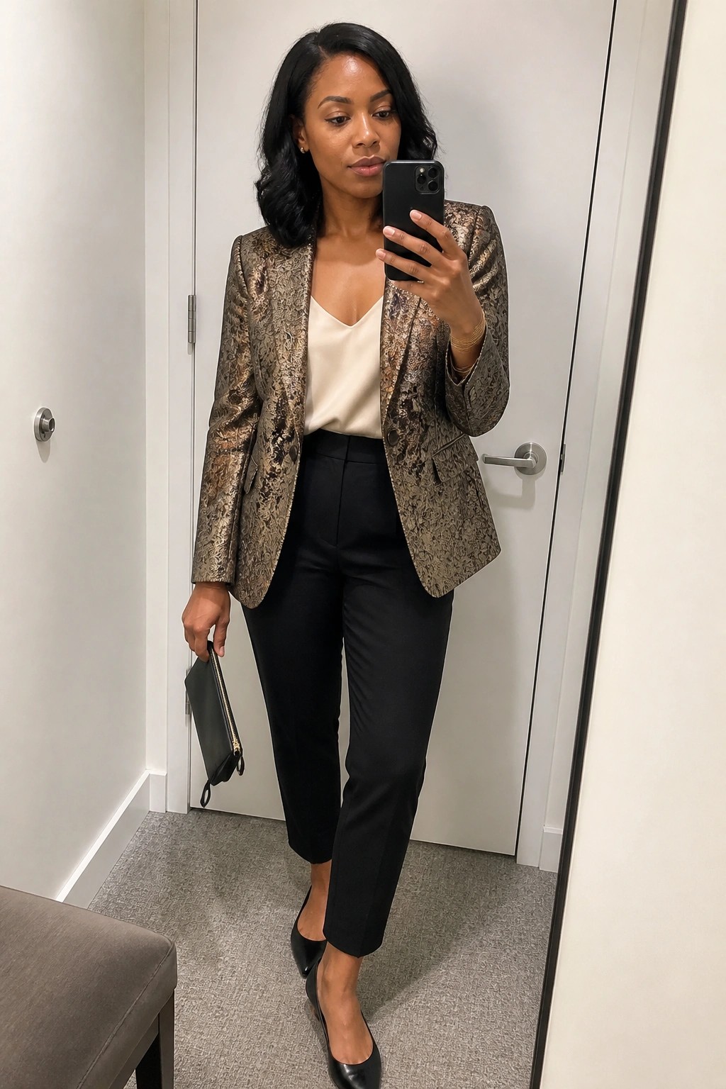 Woman in metallic patterned blazer, cream top, and black pants taking mirror selfie.
