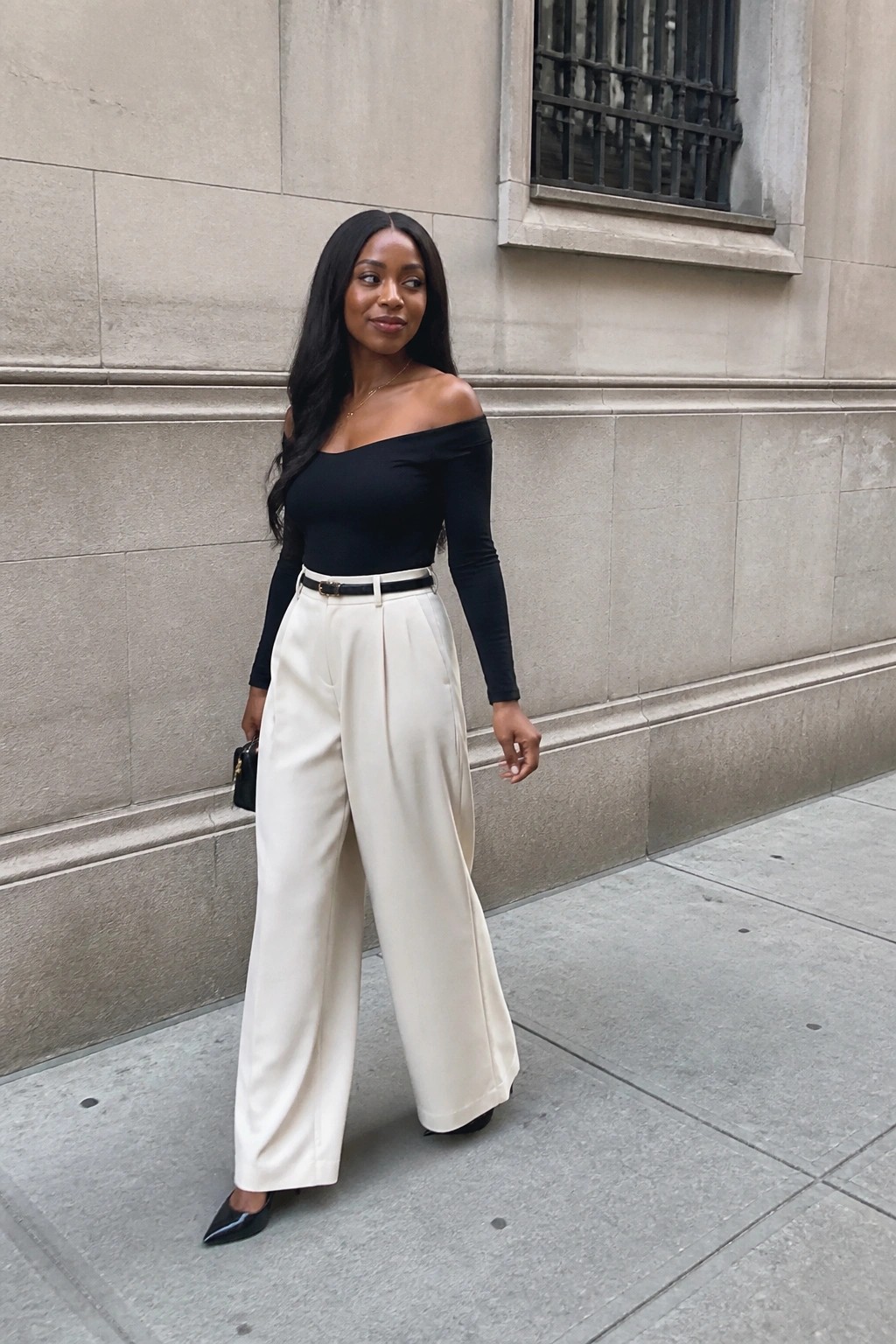 Woman wearing a black off-shoulder long sleeve bodysuit tucked into cream wide leg trousers with a black belt, black pointed heels, and holding a small black handbag.