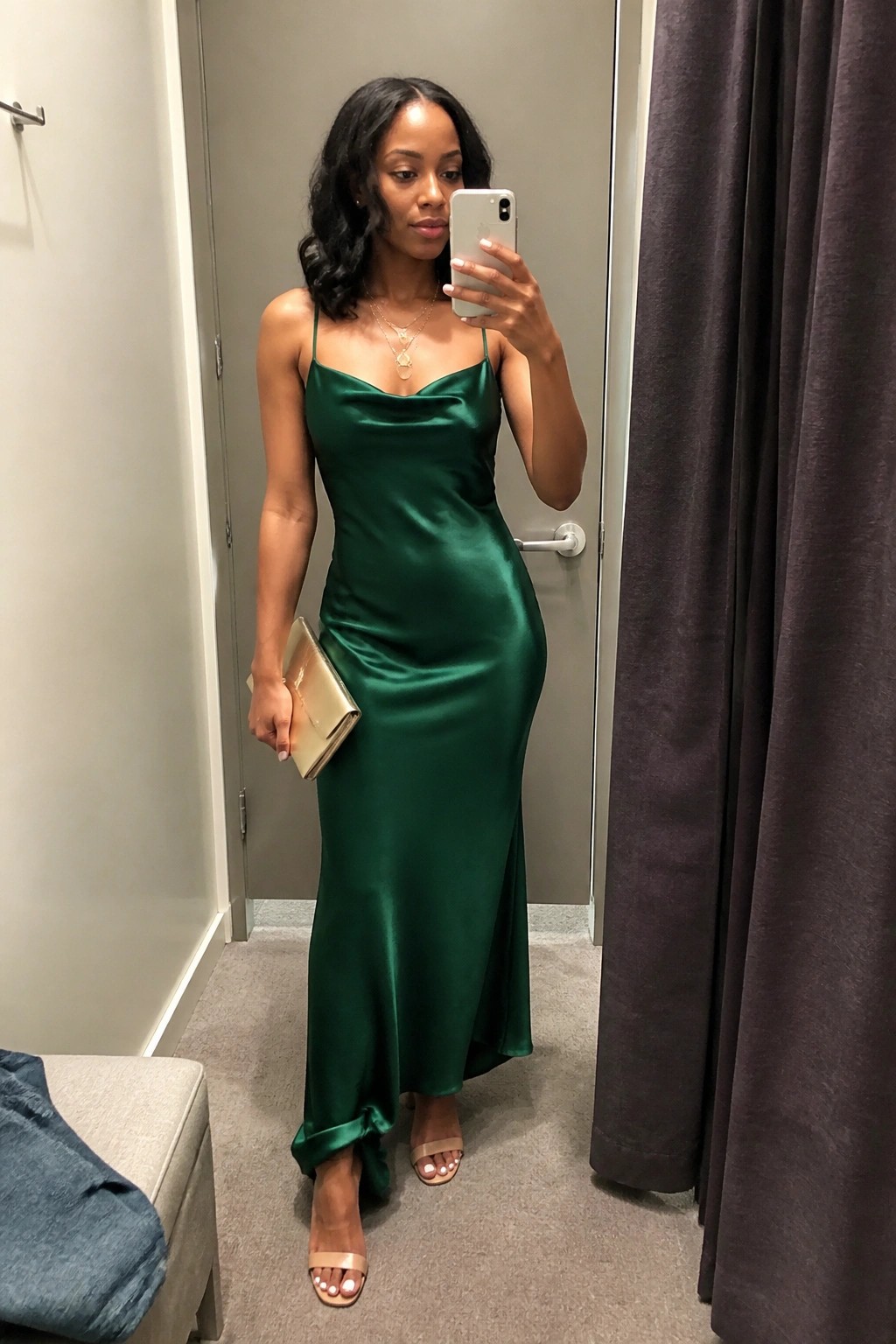 Woman wearing a long emerald green satin slip dress with thin straps and cowl neckline, holding a gold clutch and wearing layered gold necklaces with nude heels