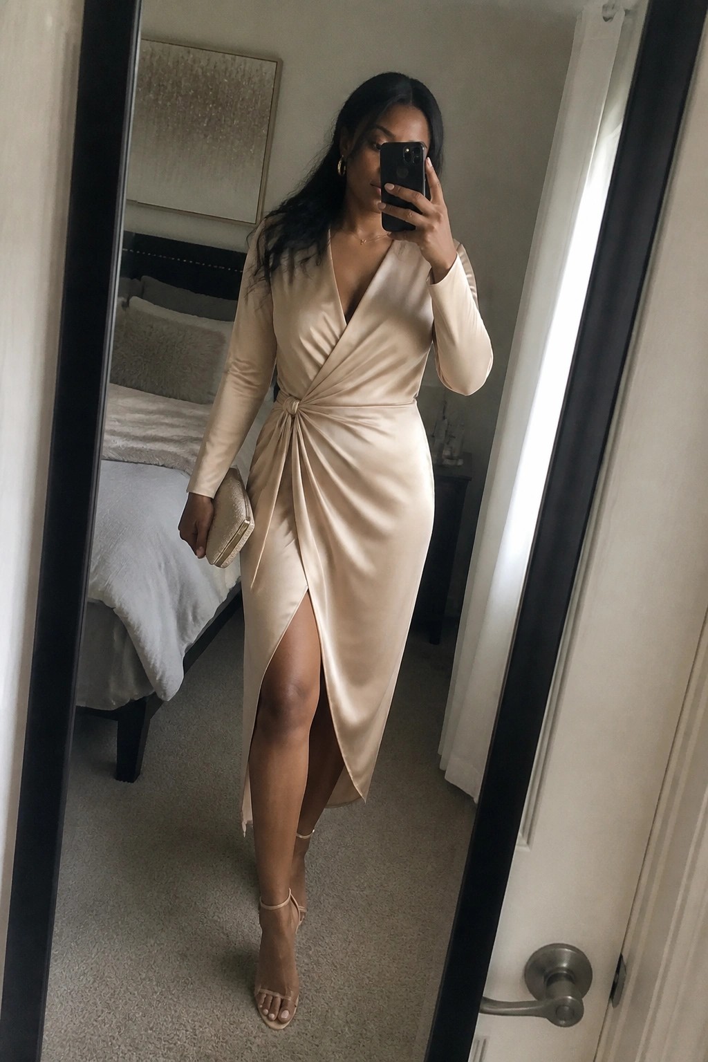 Woman in beige satin wrap dress taking mirror selfie holding phone and clutch