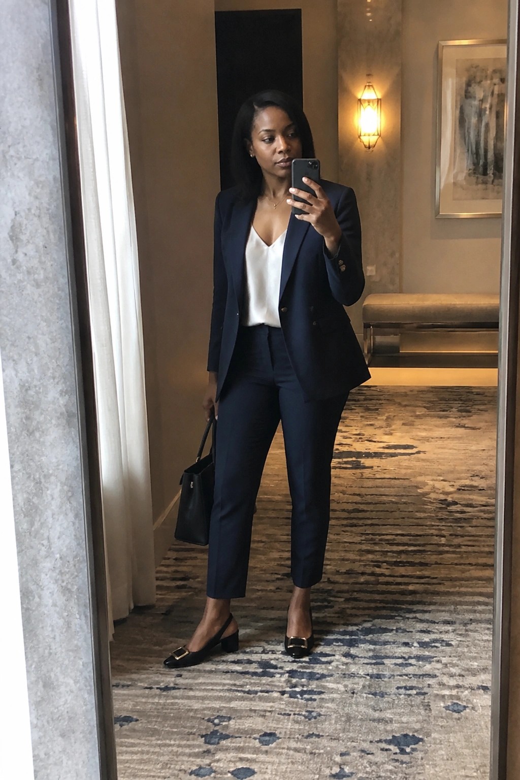 Woman in navy pantsuit taking mirror selfie while holding black handbag