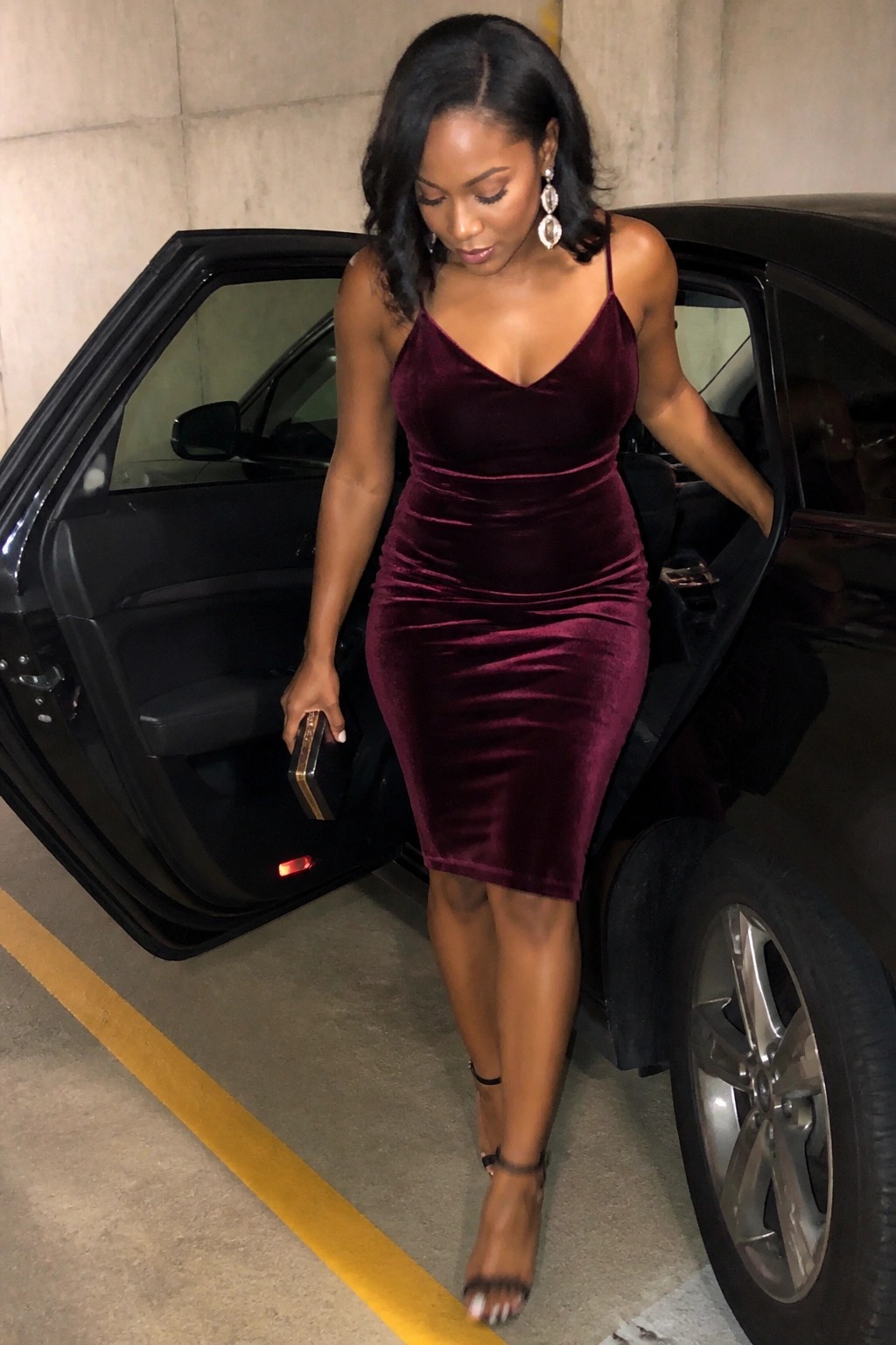 Woman in burgundy velvet dress stepping out of black car in garage.