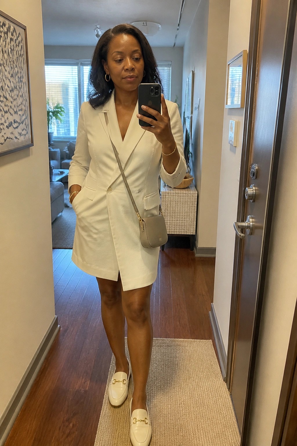Woman in white blazer dress taking mirror selfie in apartment hallway
