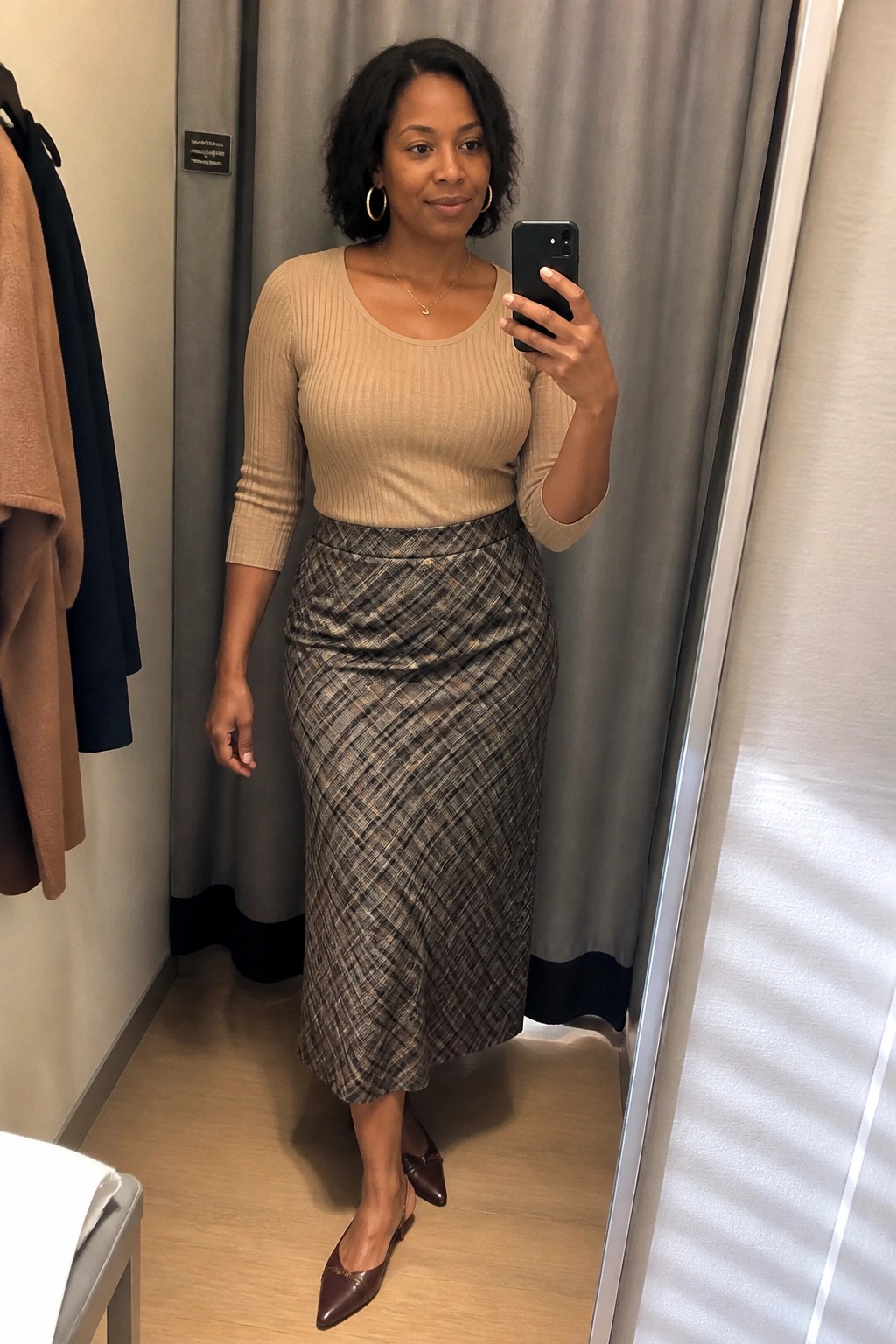Woman wearing a beige ribbed knit top tucked into a brown and black plaid midi skirt with brown pointed toe heels and gold hoop earrings while taking a mirror selfie.