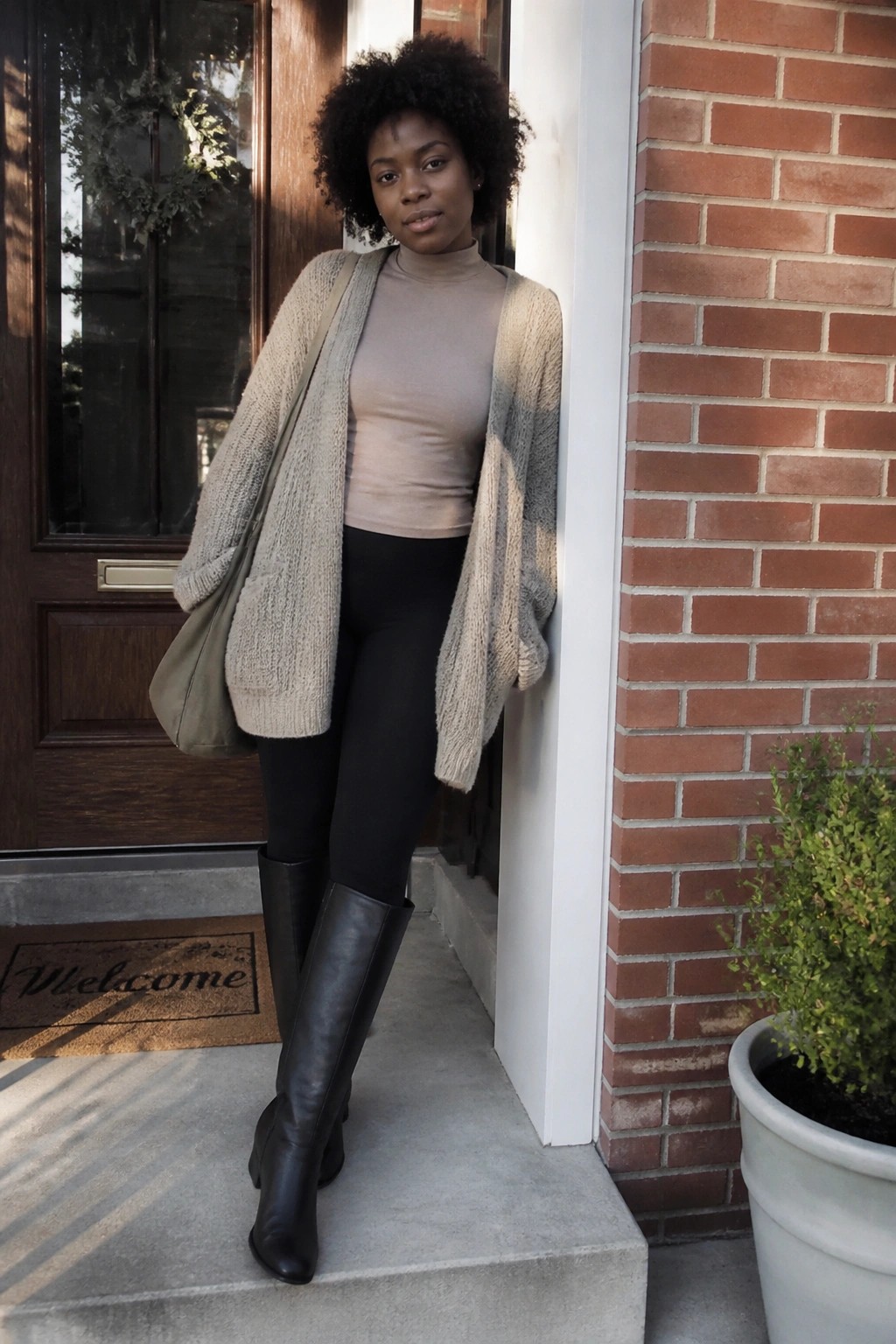 Woman in beige turtleneck, gray cardigan, black leggings and boots at doorway