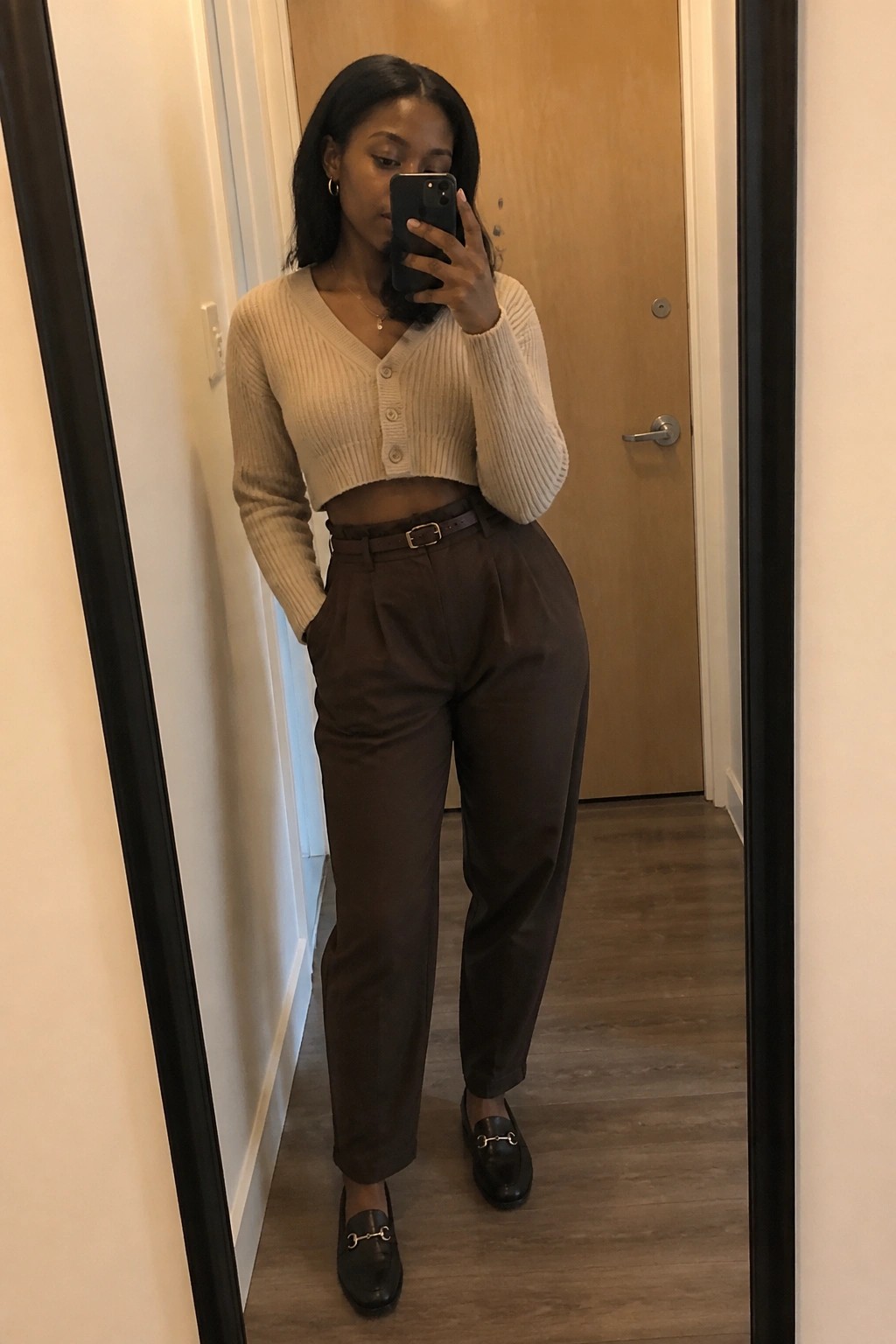 Woman in beige cropped cardigan and brown trousers taking mirror selfie