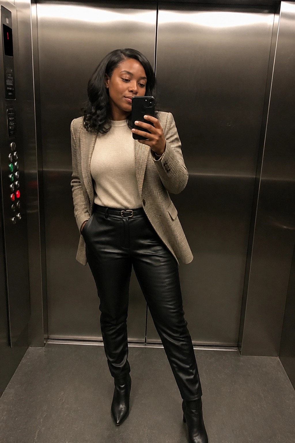 Woman standing in an elevator wearing a beige plaid blazer over a cream ribbed sweater, black leather pants with a gold buckle belt, and black pointed ankle boots while holding a phone for a mirror selfie.