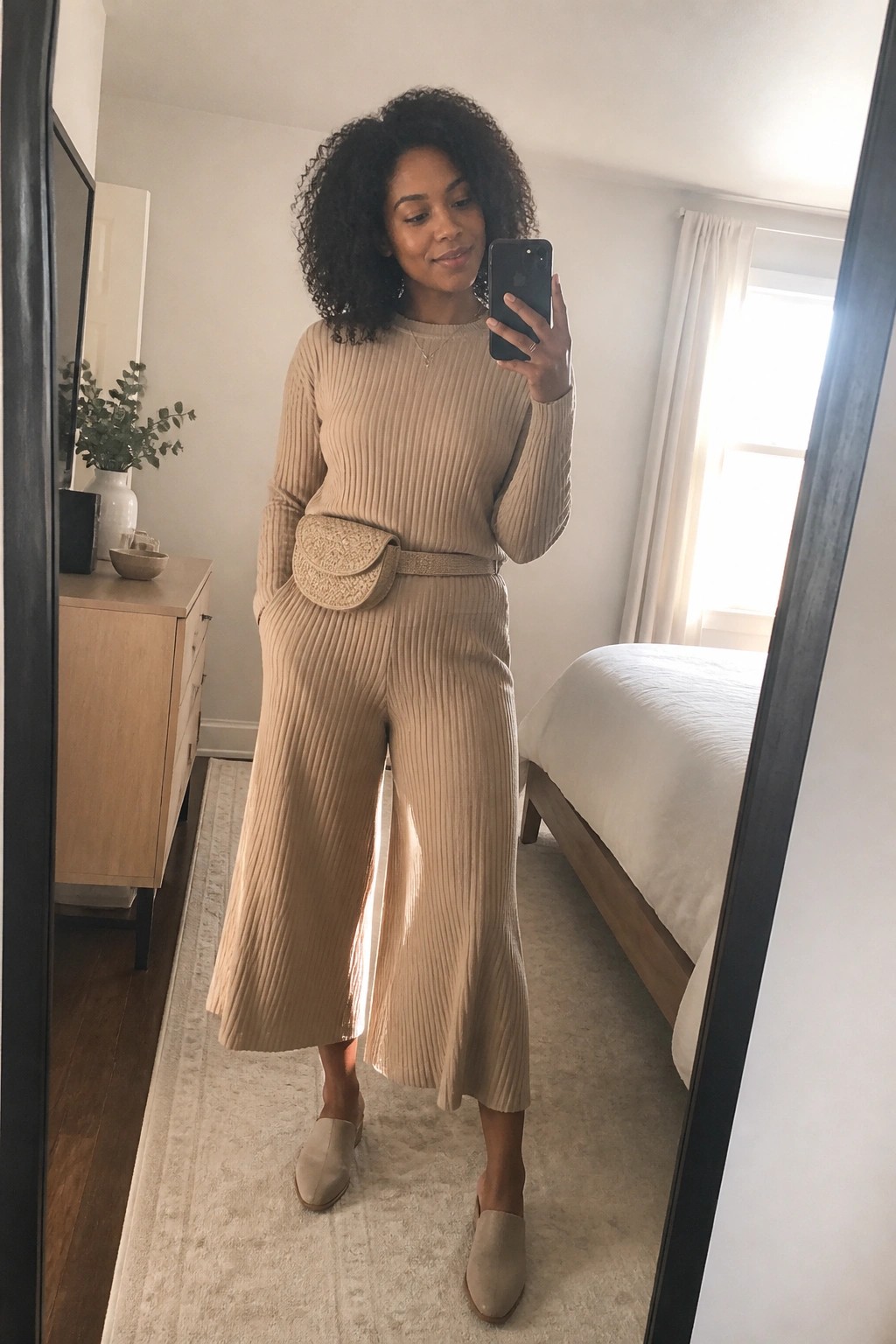 A woman wearing a beige ribbed knit sweater and matching wide leg cropped pants with a woven belt bag, layered necklaces, and beige mules, posing in a bedroom mirror selfie.