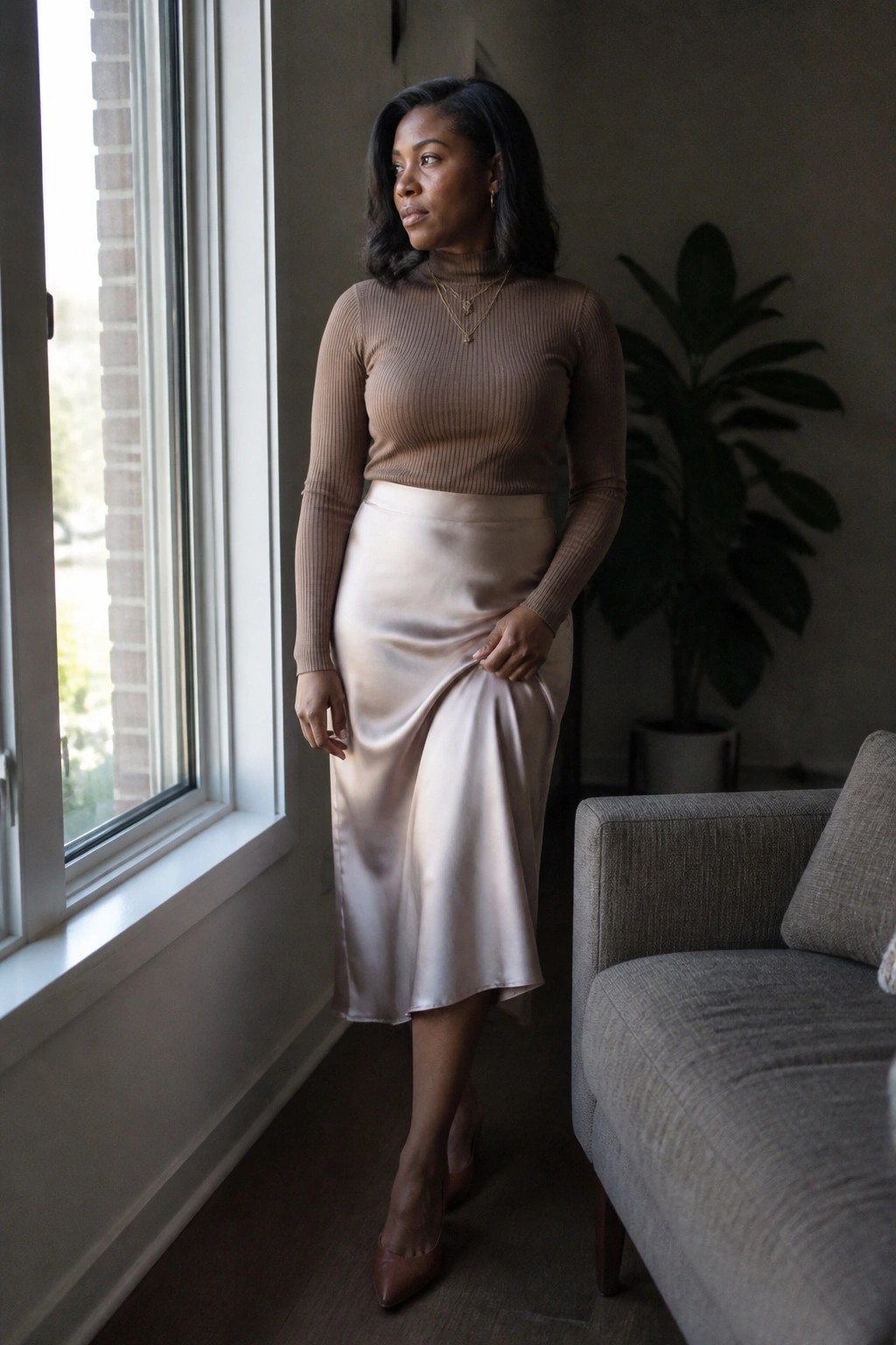 Woman in brown ribbed turtleneck sweater paired with beige satin midi skirt, brown pointed heels, and layered gold necklaces standing indoors near a window.