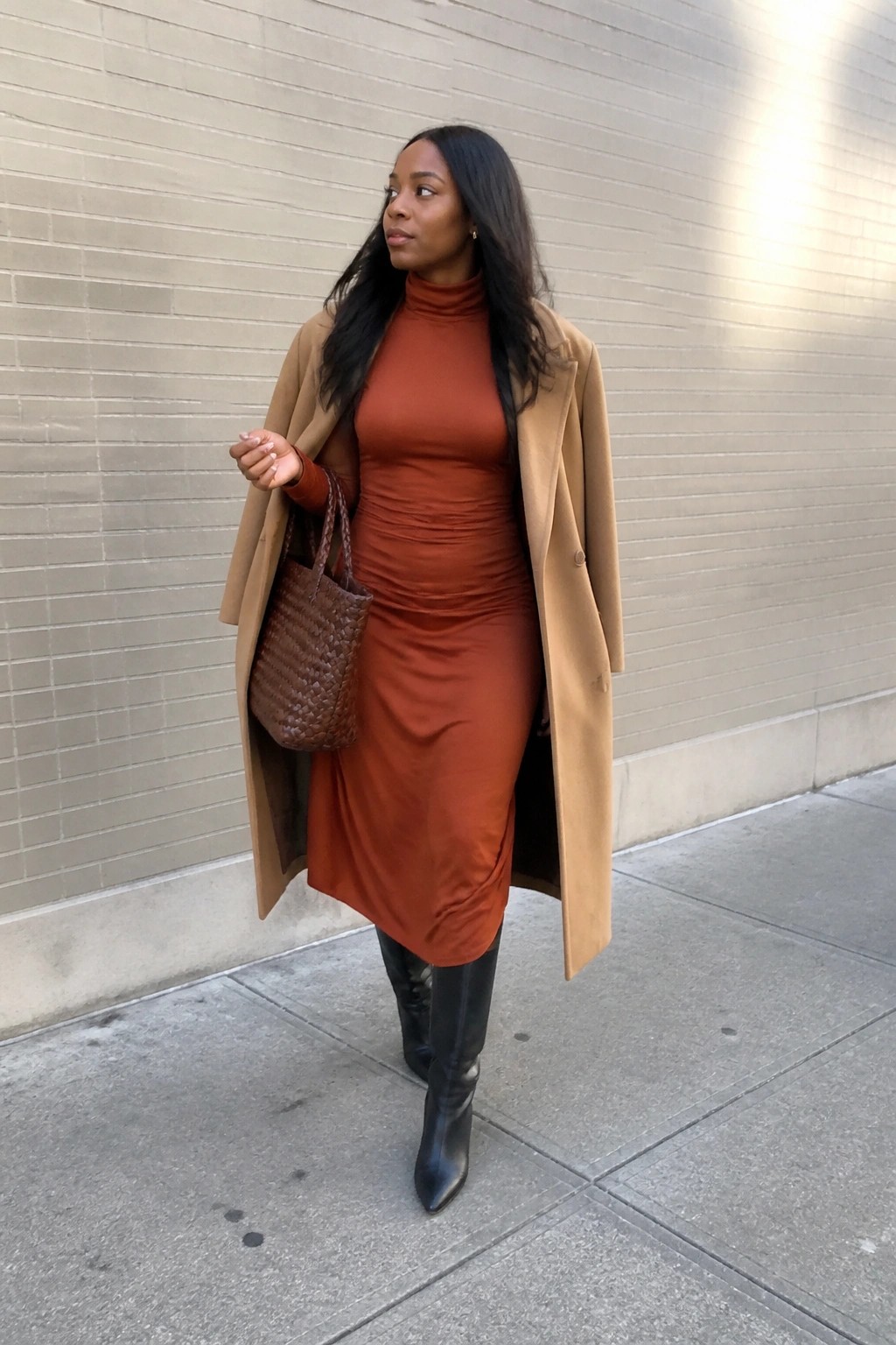 Woman wearing a rust orange ruched turtleneck midi dress under an open camel long coat with black knee high pointed boots and carrying a brown woven leather tote bag.
