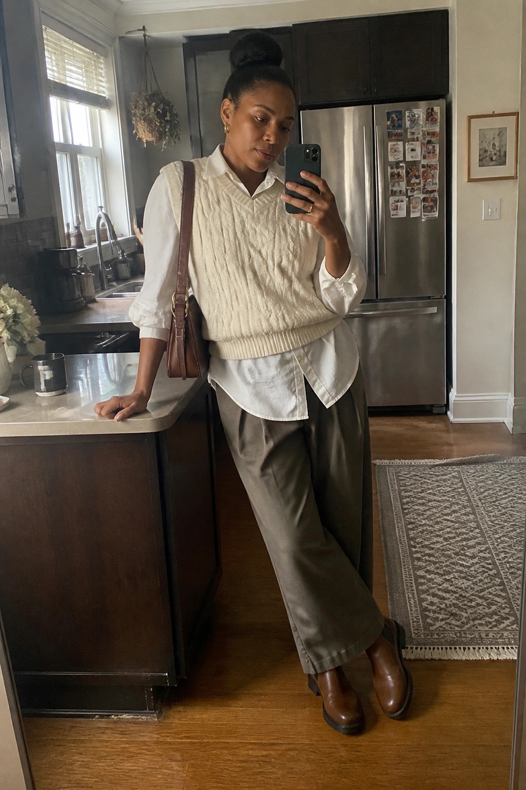 Black woman wearing a white button-down shirt layered under a cream cable-knit sweater vest, paired with olive wide-leg trousers and brown leather boots, holding a phone in a kitchen setting.