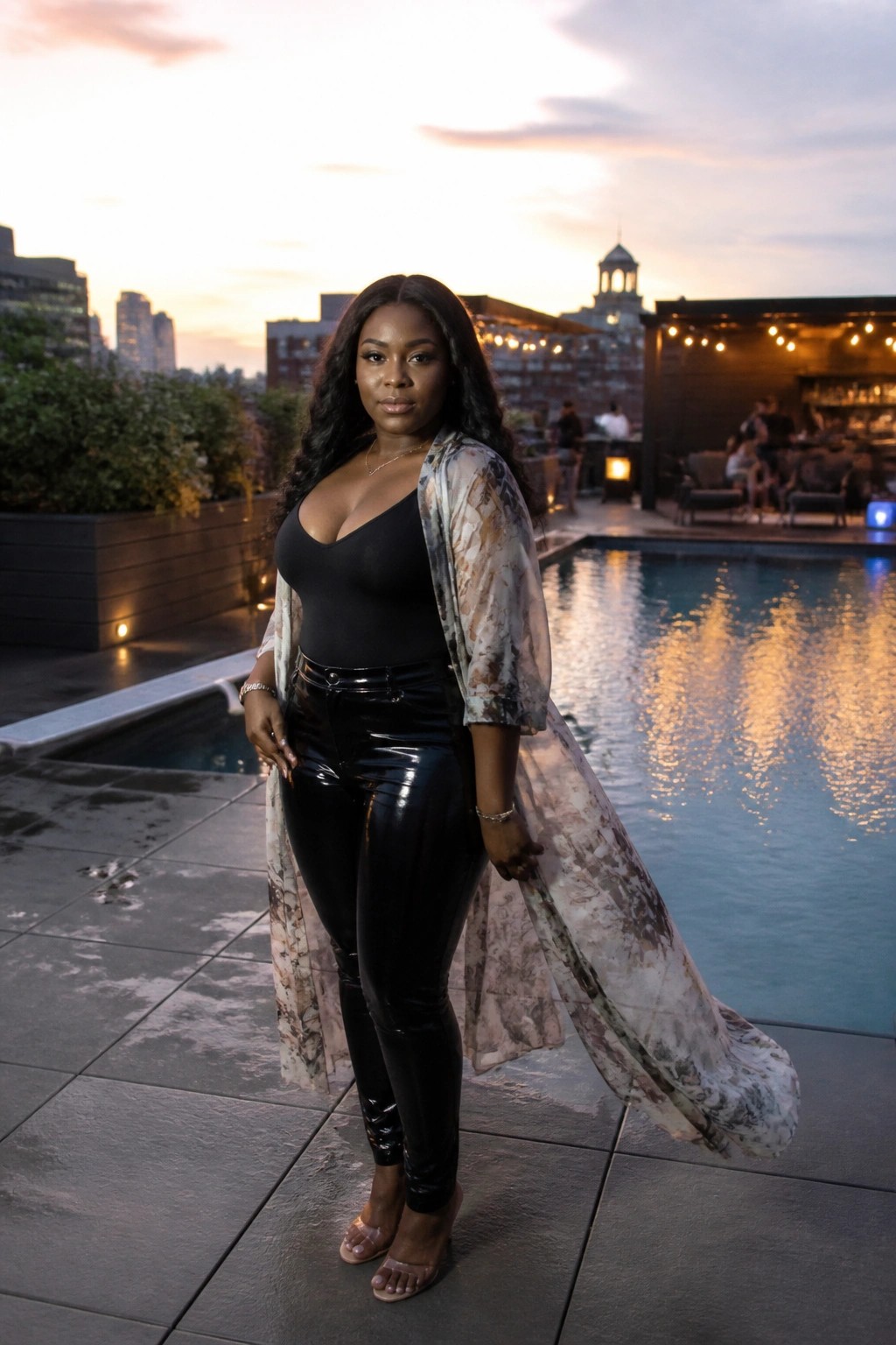 Woman in black bodysuit and shiny pants with sheer patterned robe by pool at sunset