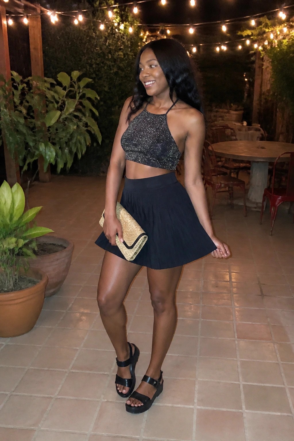 Woman in sparkly black halter crop top and pleated skirt holding gold clutch on patio