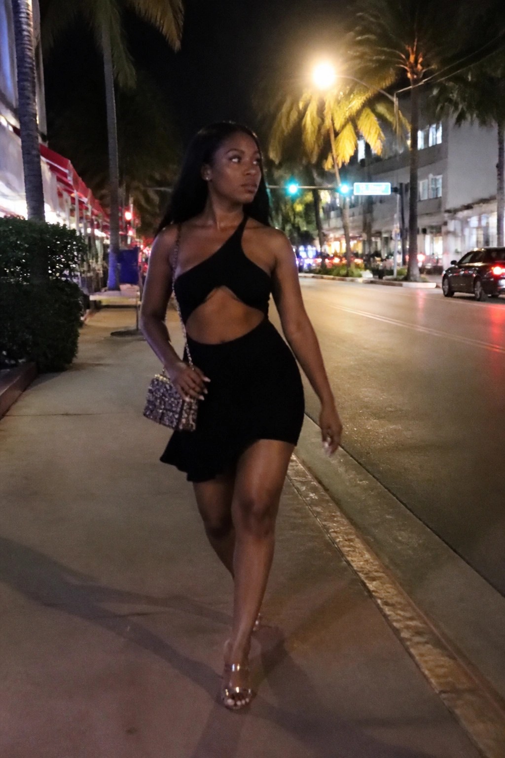 Woman in black cut-out dress walks nighttime palm-lined city street with sparkly purse