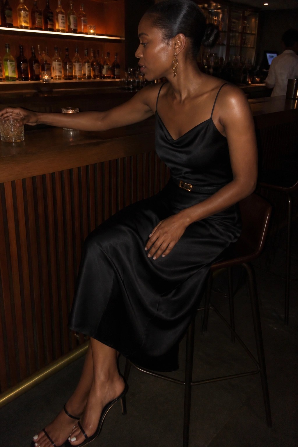 Woman in black satin dress seated at wooden bar holding drink.