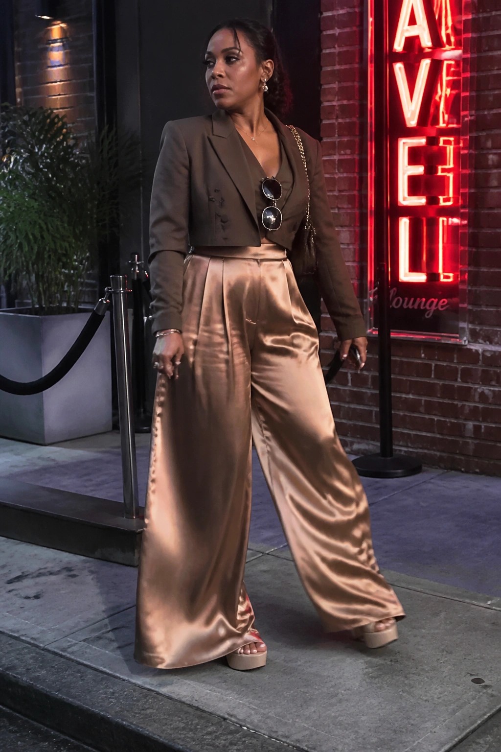 Woman in brown cropped blazer and shiny satin wide-leg pants on nighttime sidewalk.