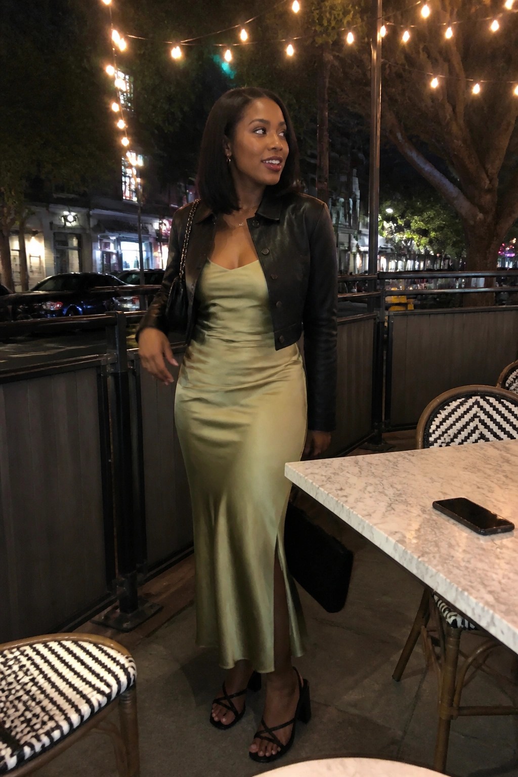 Woman in sage satin slip dress and black leather jacket at night cafe table