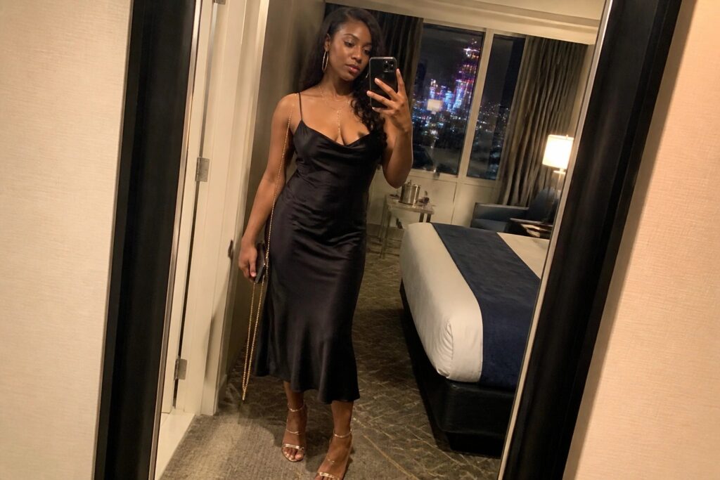 Woman in long black satin dress taking mirror selfie in hotel room at night.