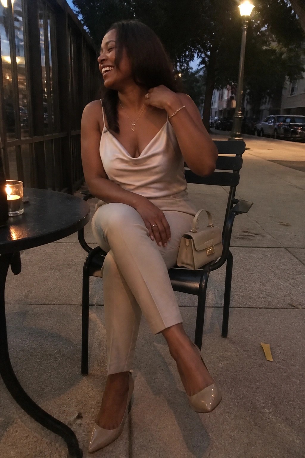 Woman in beige satin camisole and pants seated on black chair outdoors at night