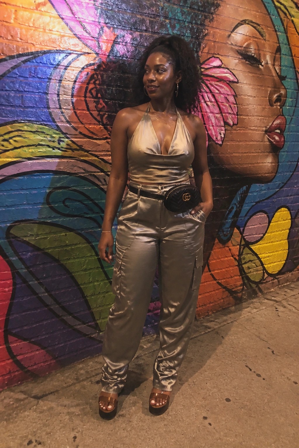 Woman in silver satin halter top and cargo pants before colorful brick mural with Gucci bag