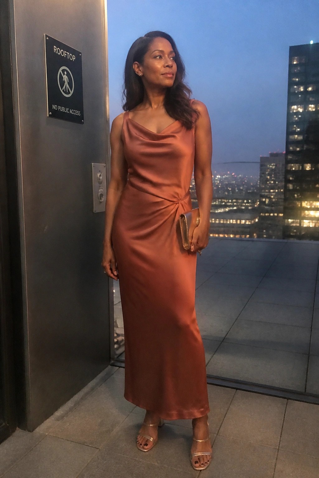 Woman in terracotta satin dress posing on city rooftop at dusk near door