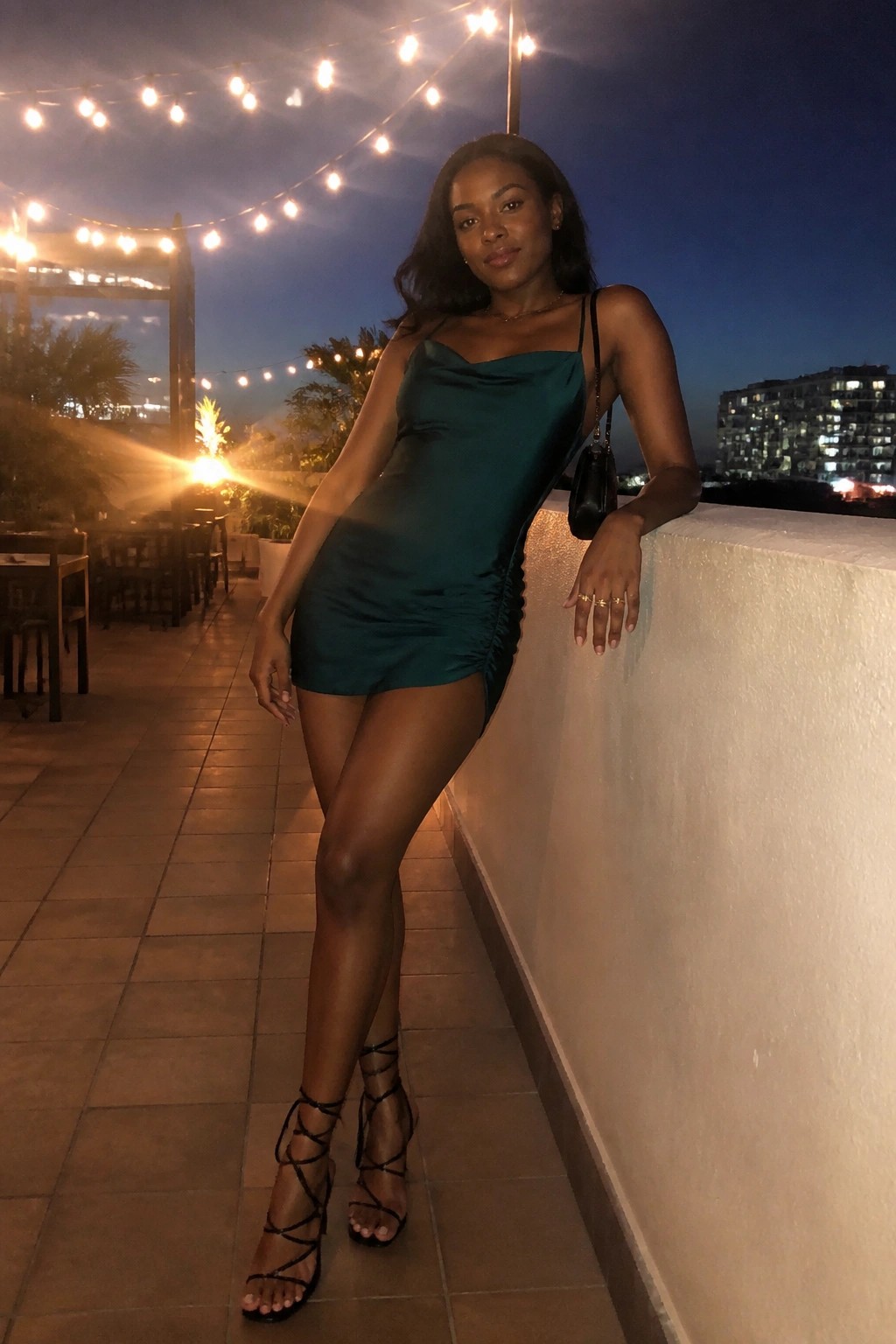 Woman in green satin mini dress with strappy heels on night balcony under lights