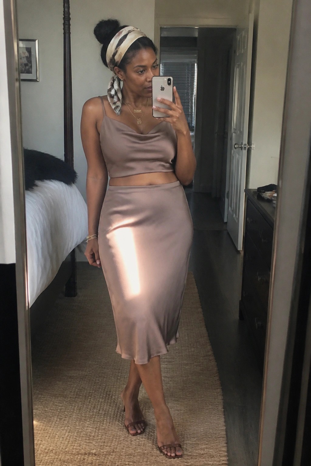 Woman in light brown satin crop top and skirt taking bedroom mirror selfie