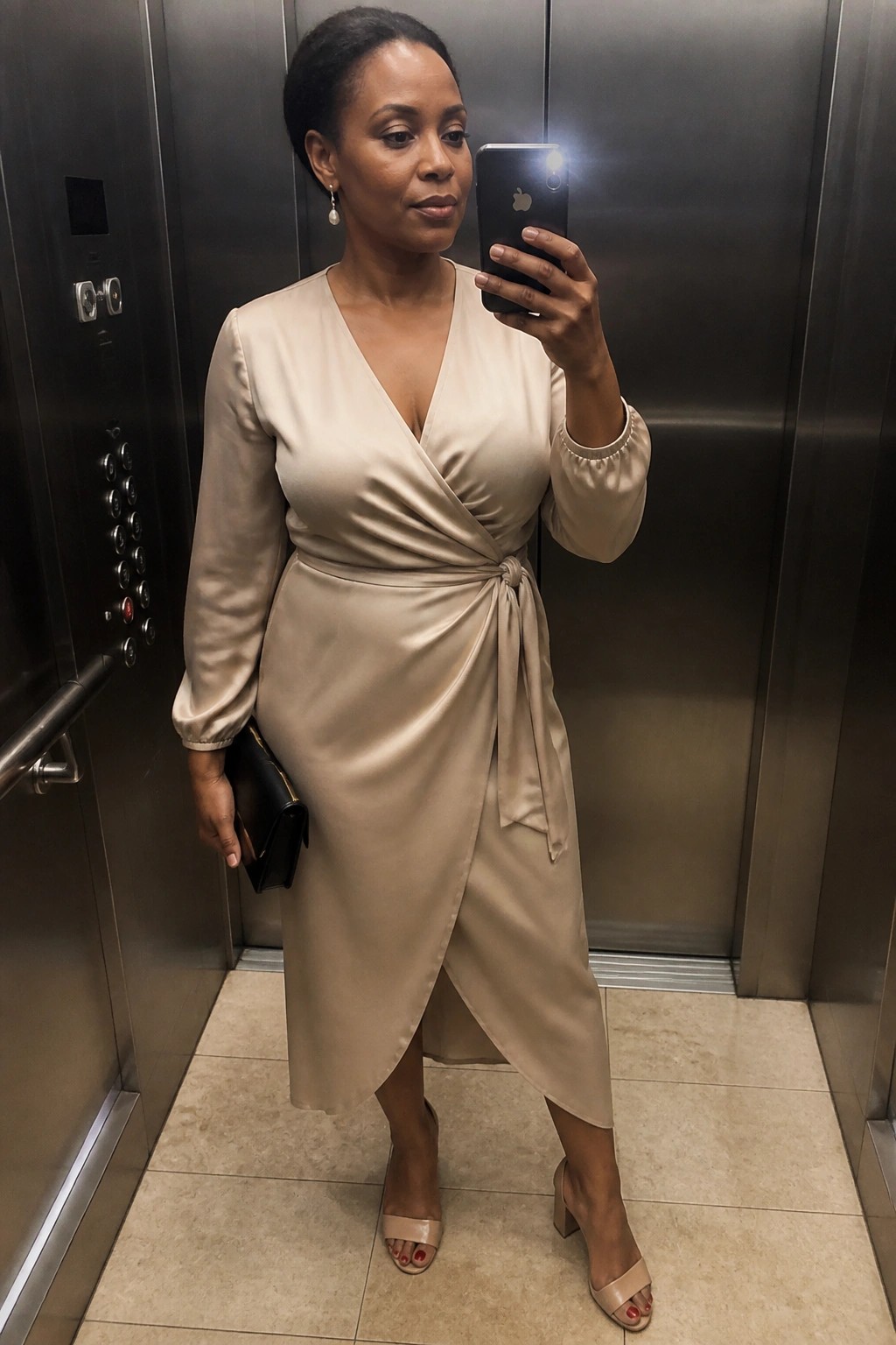Woman in beige wrap dress taking selfie inside metallic elevator with black clutch