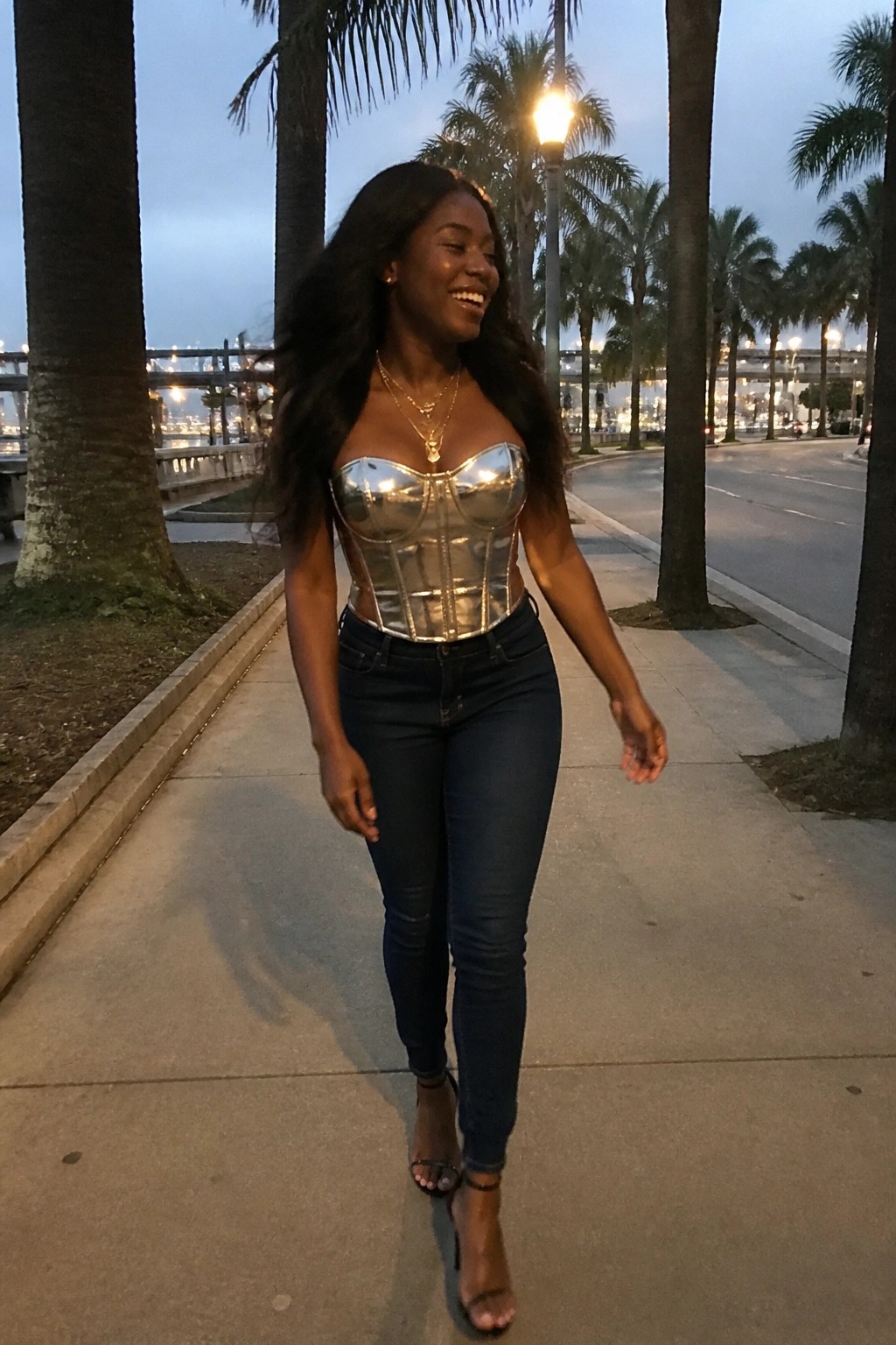Woman in silver corset and jeans walking on palm-lined sidewalk at dusk