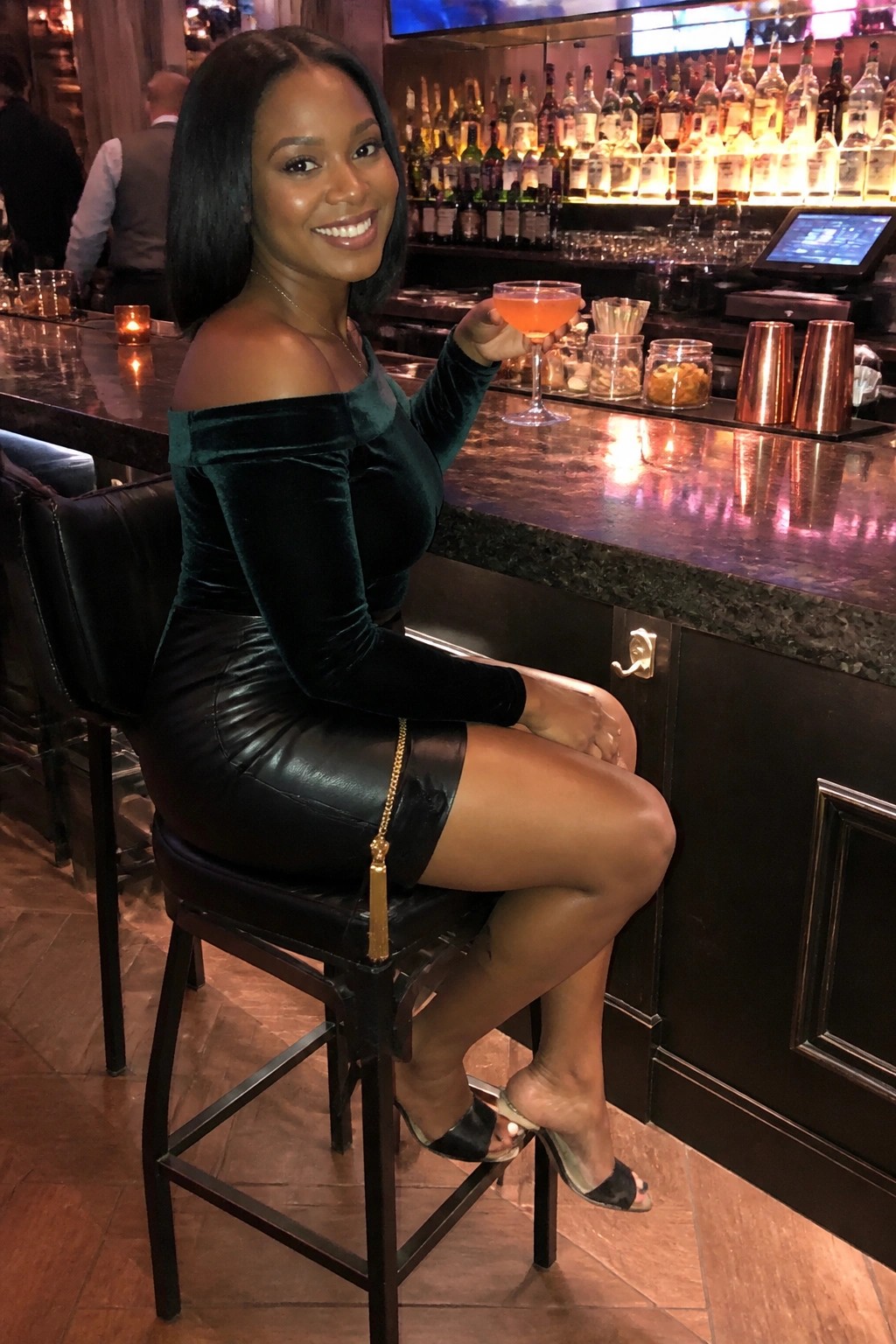 Smiling woman in green velvet top and black skirt holds pink cocktail at bar.