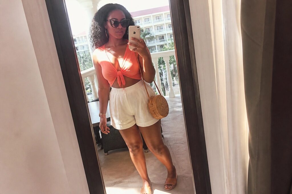 21 Miami Spring Break Outfits for Black Women Built for Beach Parties Woman in orange knotted top and white shorts taking balcony selfie overlooking ocean