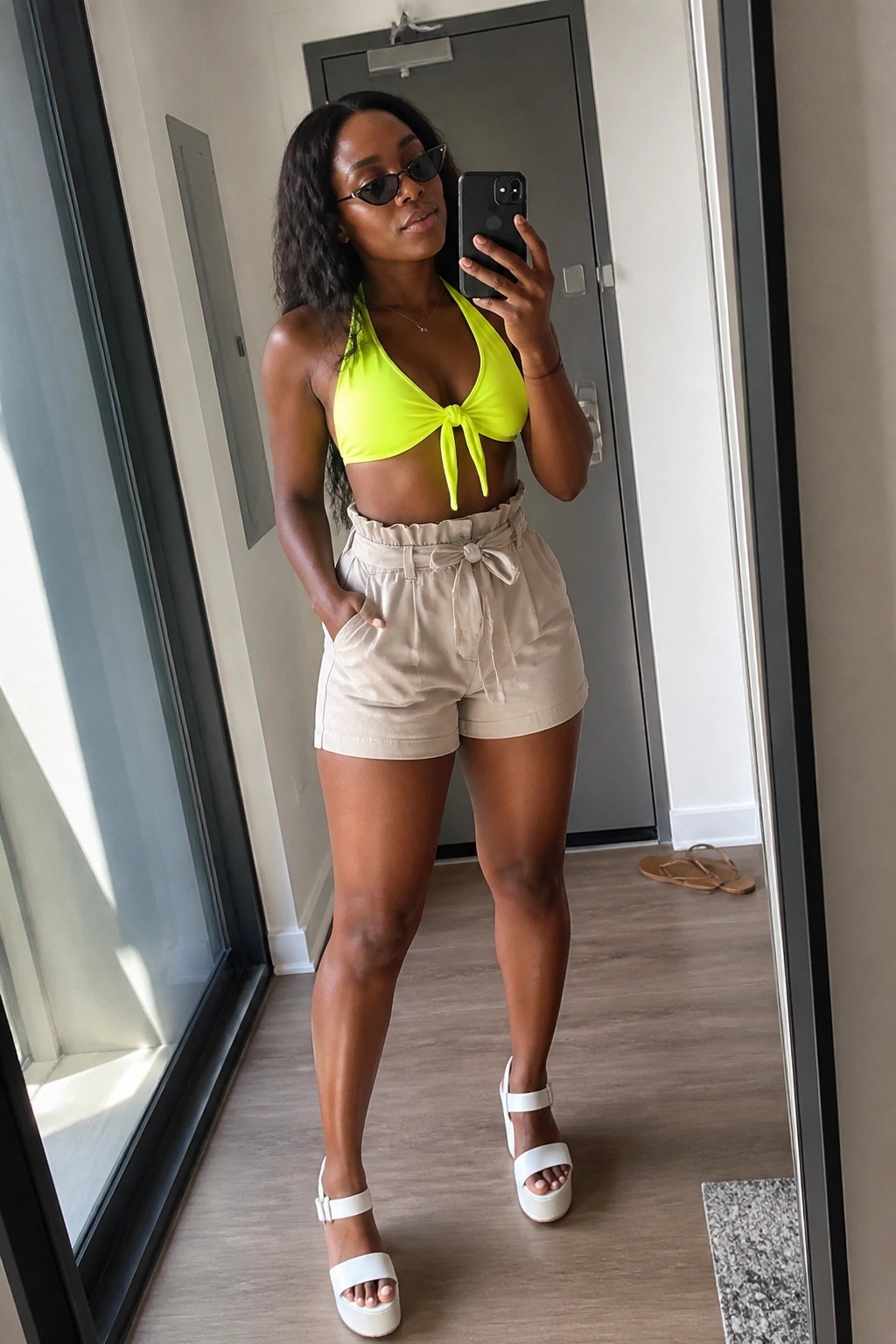 Woman in mirror selfie wearing neon yellow halter crop top tied at center, beige high waisted paperbag shorts with tie belt, and white platform sandals.