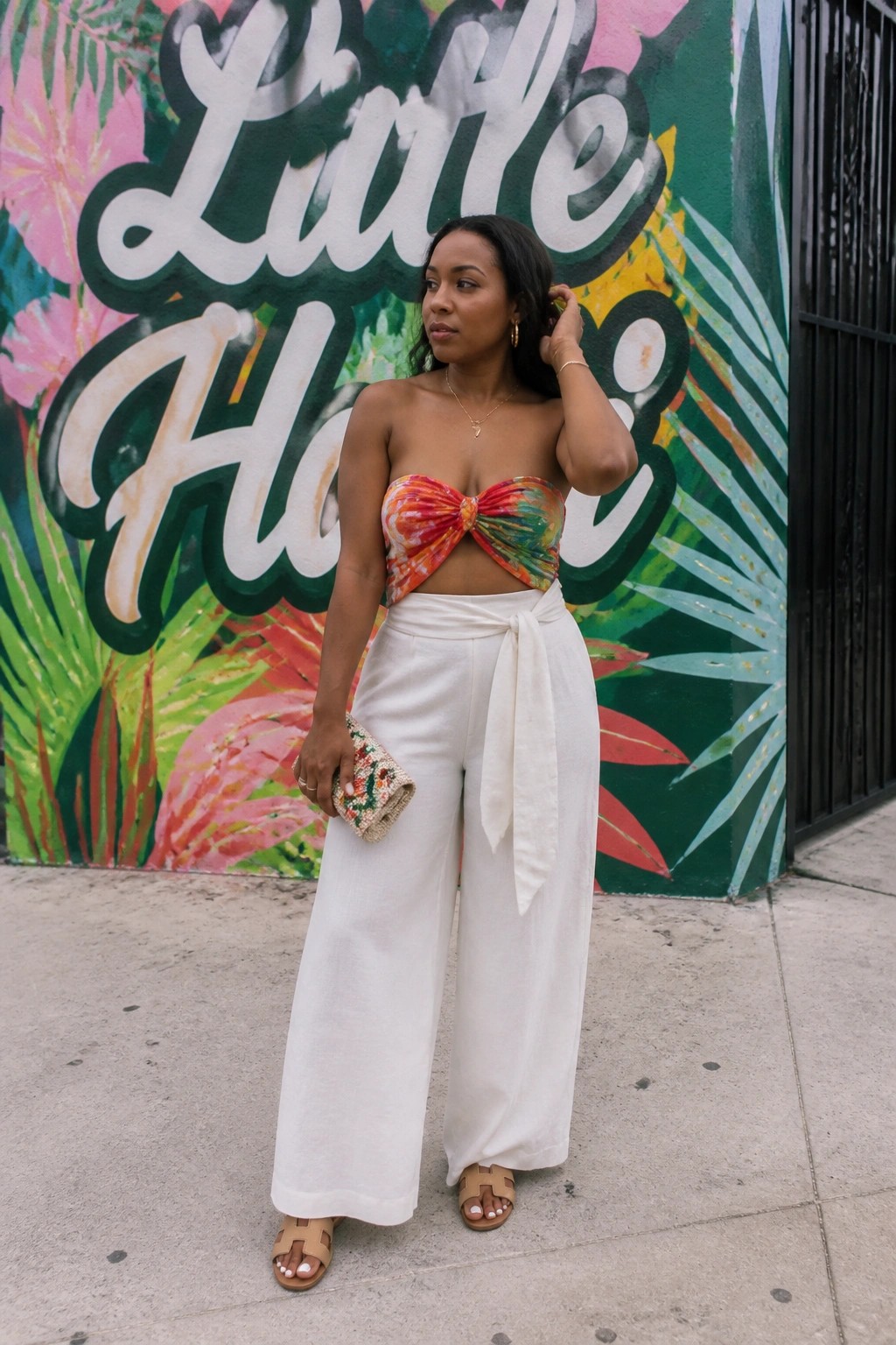 Woman in colorful tie-dye strapless crop top with front knot, white wide-leg pants with waist tie, beige flat sandals, and beaded clutch standing in front of mural.