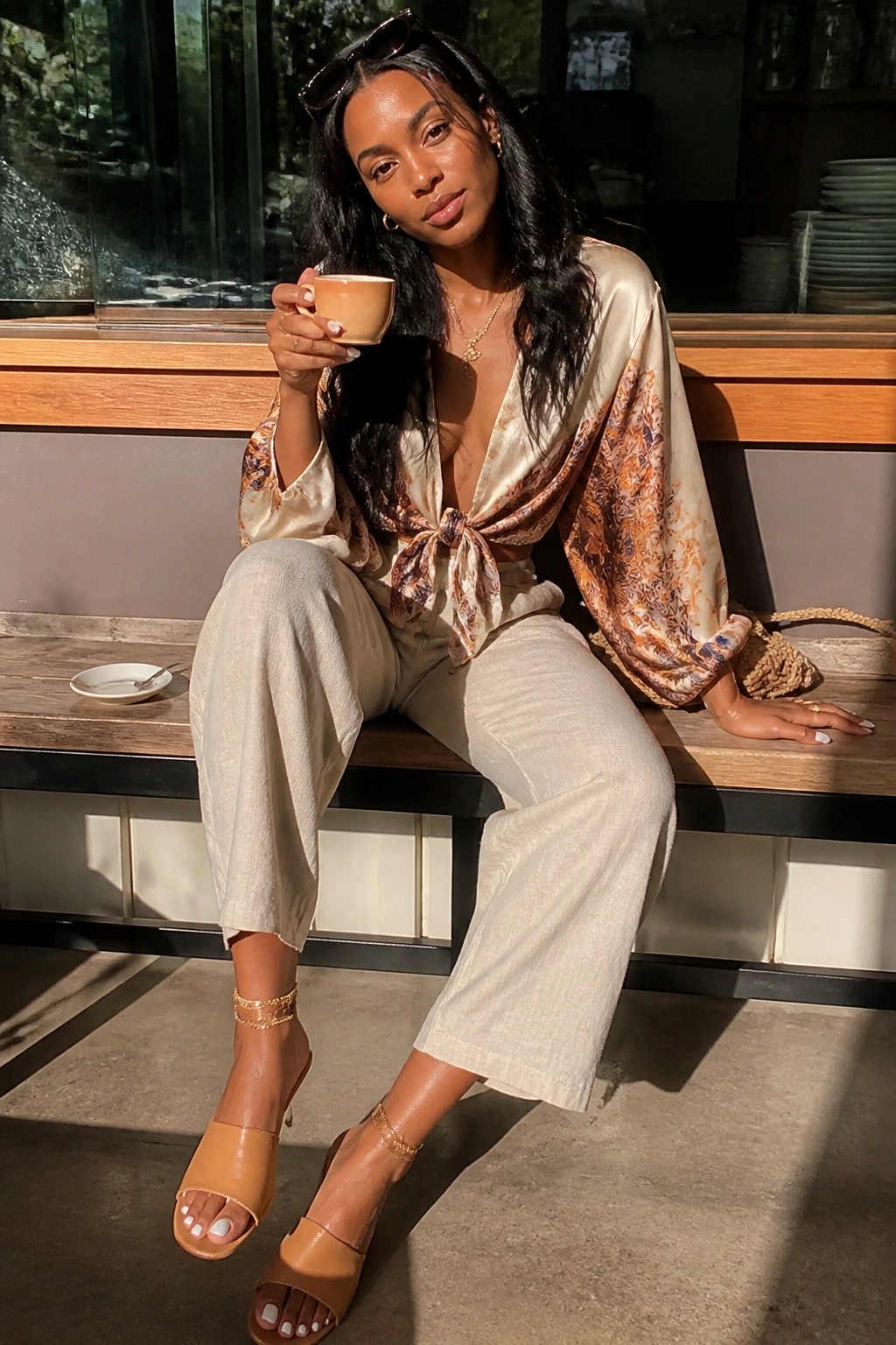 Woman in patterned satin top and wide beige pants holding coffee on bench.
