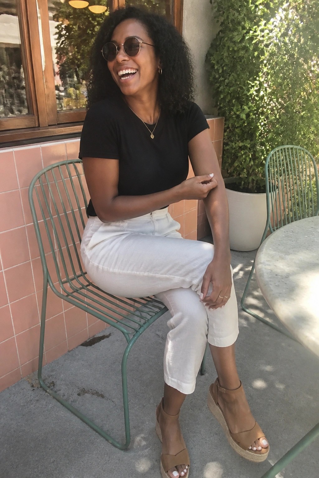 Woman in black short sleeve t-shirt, white cropped pants, and brown wedge sandals seated on a green metal chair with gold necklace and sunglasses.