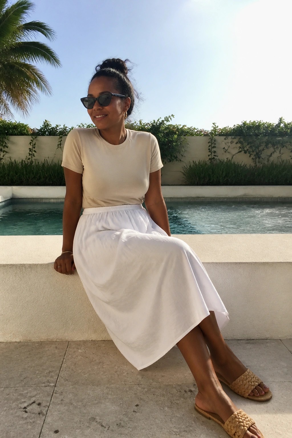 Woman in cream top and white skirt seated poolside by turquoise water