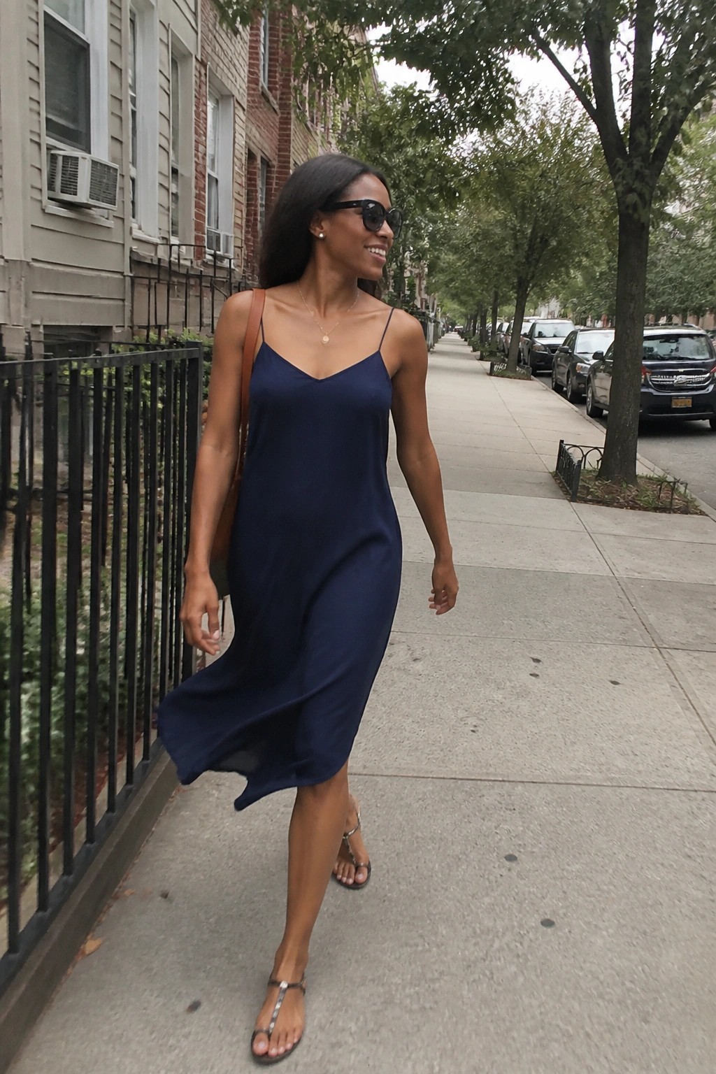 Woman in navy slip dress walking on tree-lined urban sidewalk with parked cars
