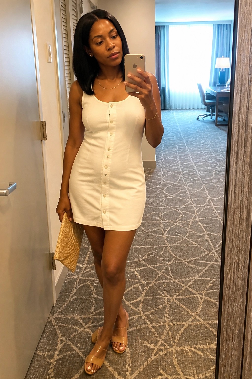 Woman in white button-front mini dress taking mirror selfie in hotel room.