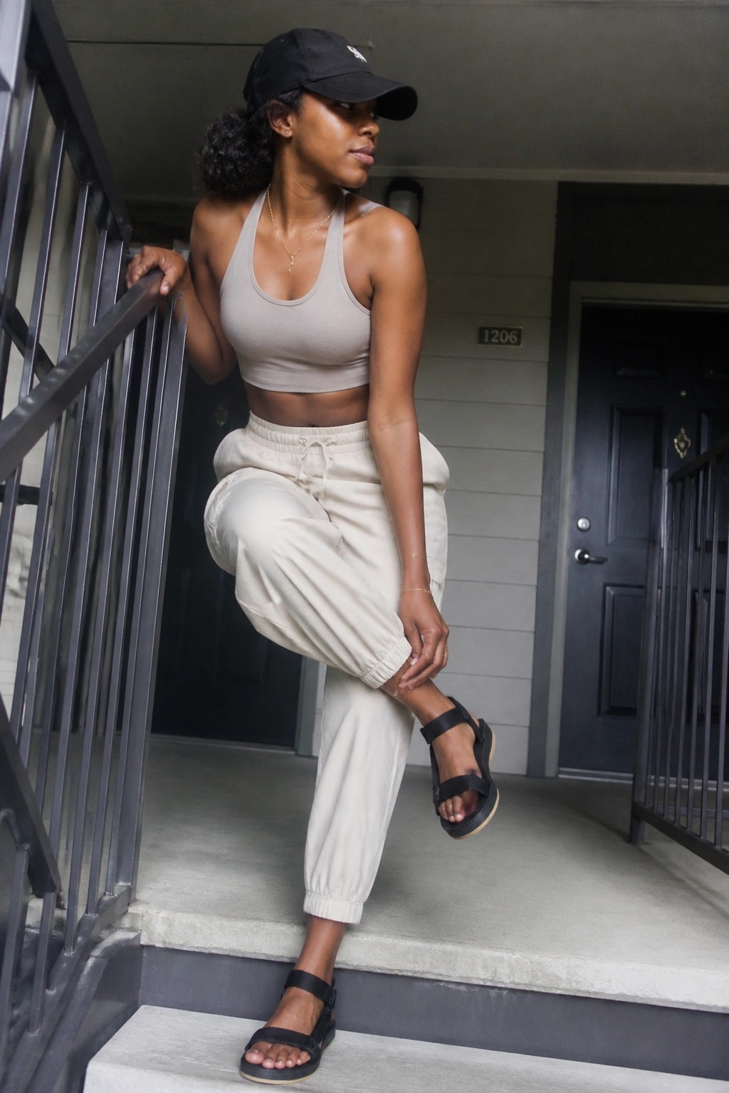 Woman in beige crop top and joggers posing on building stairs with black cap and sandals.