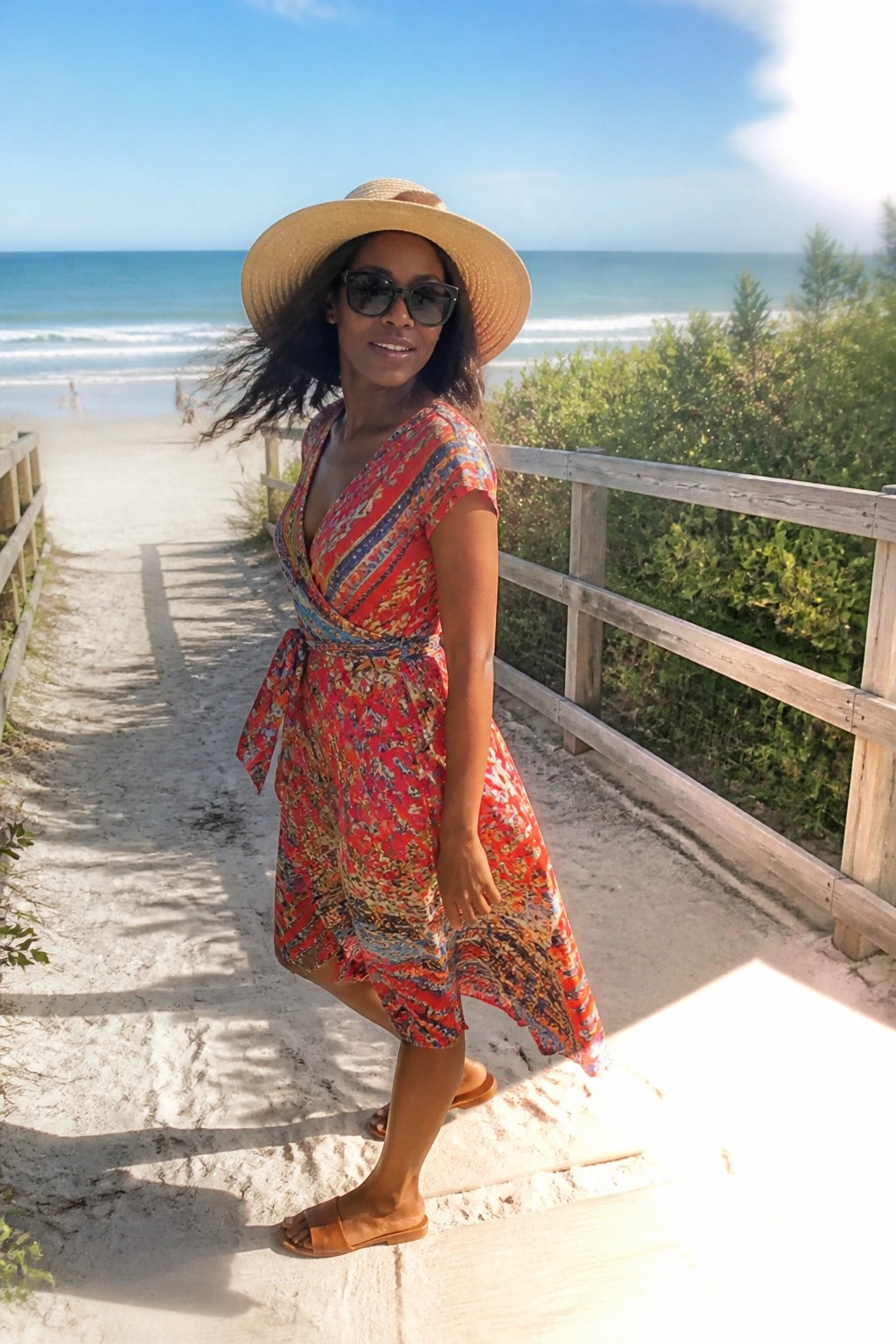 Woman in red patterned wrap dress and straw hat on sunny beach steps.