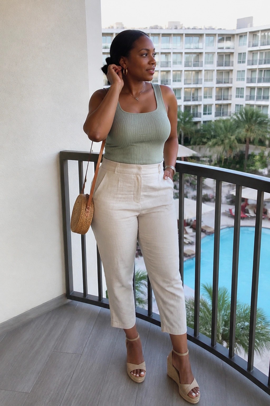 Woman in sage green top and beige pants on resort balcony by pool.