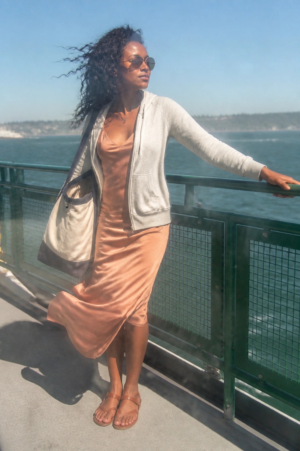 Woman wearing a peach satin slip midi dress layered under an open light gray zip hoodie with brown flat sandals and a beige tote bag standing by a green railing.