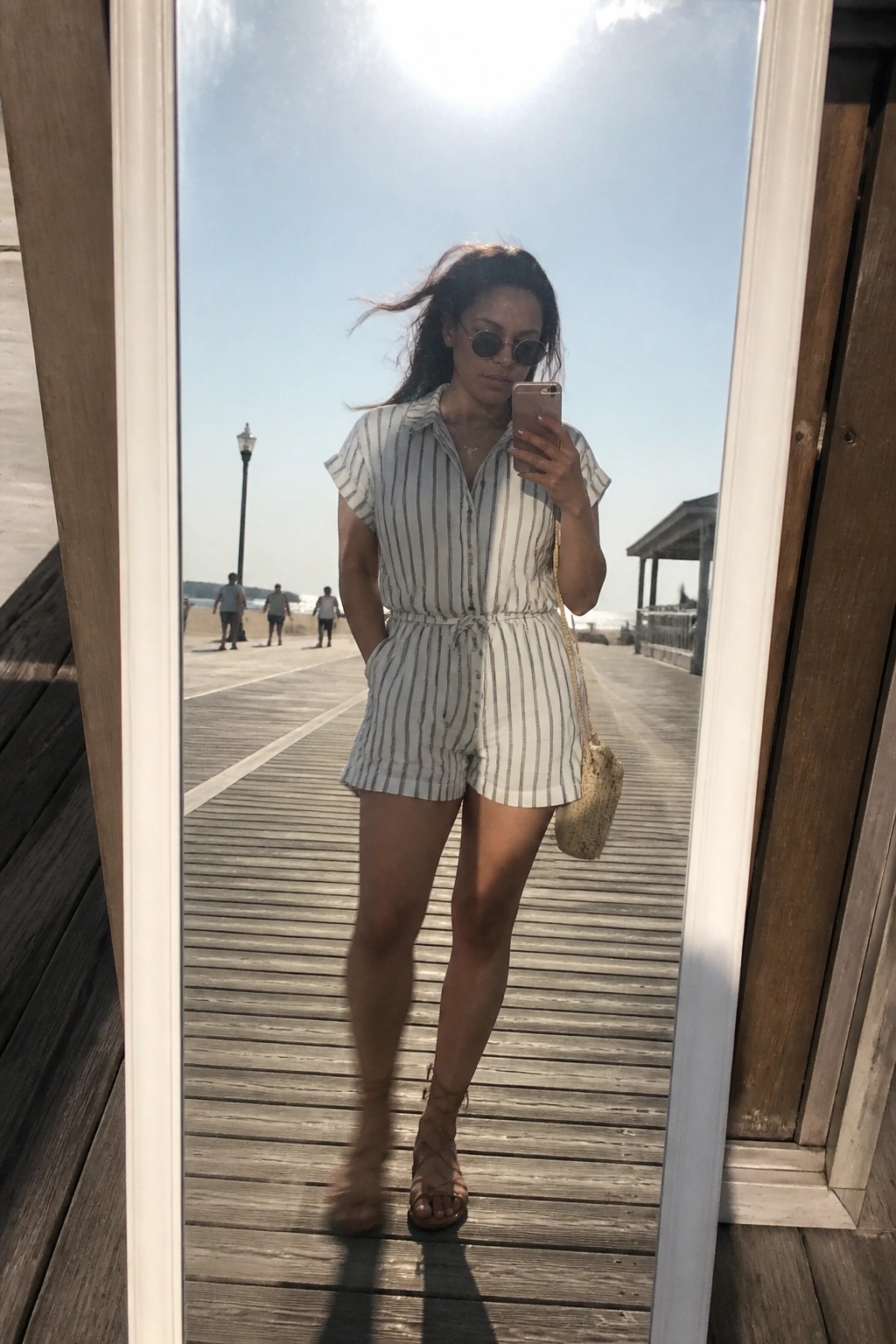Woman in striped romper taking mirror selfie on sunny ocean boardwalk